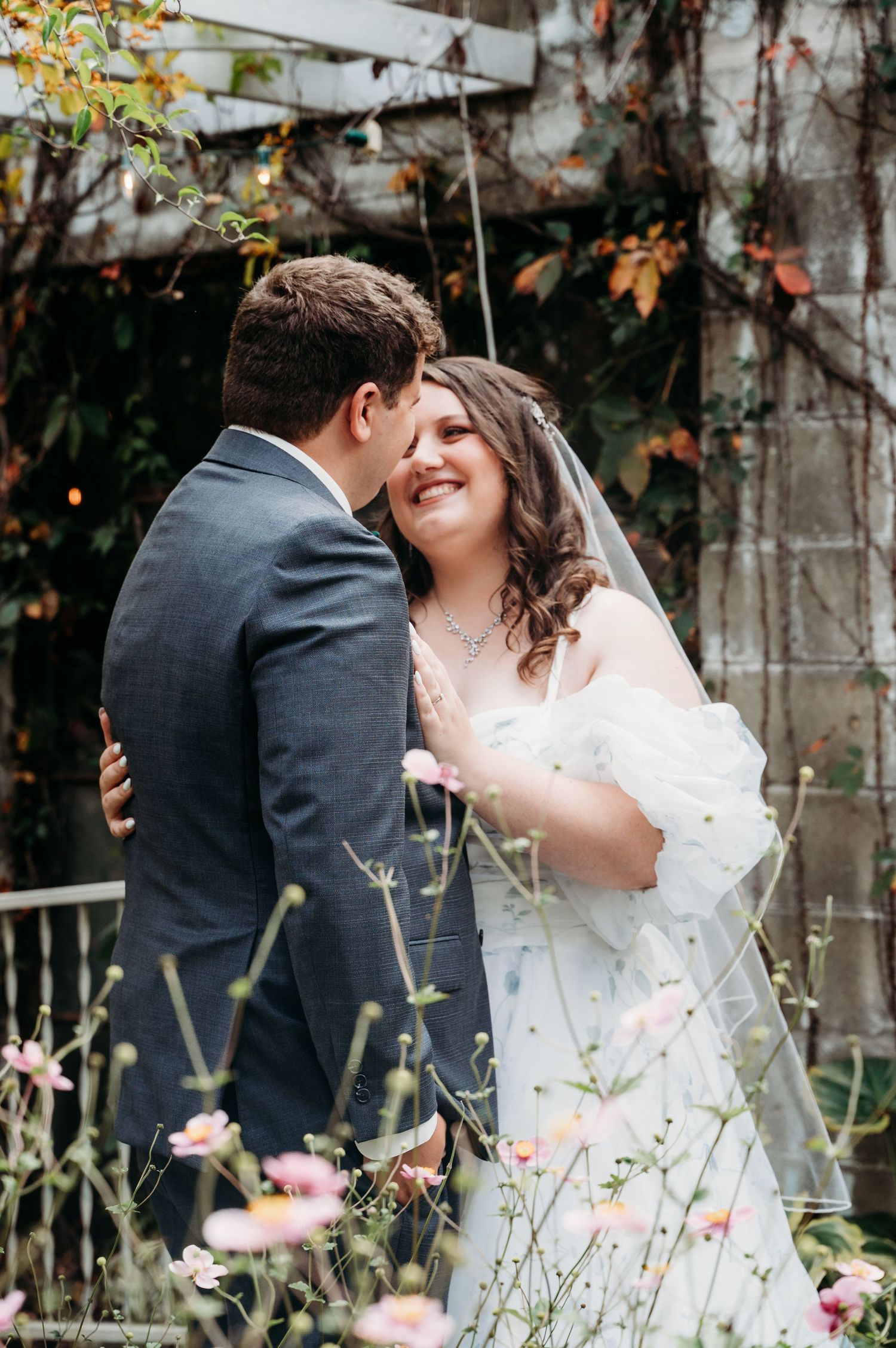 A romantic wedding couple embraces in a garden setting with soft autumn foliage and delicate flowers in the background.