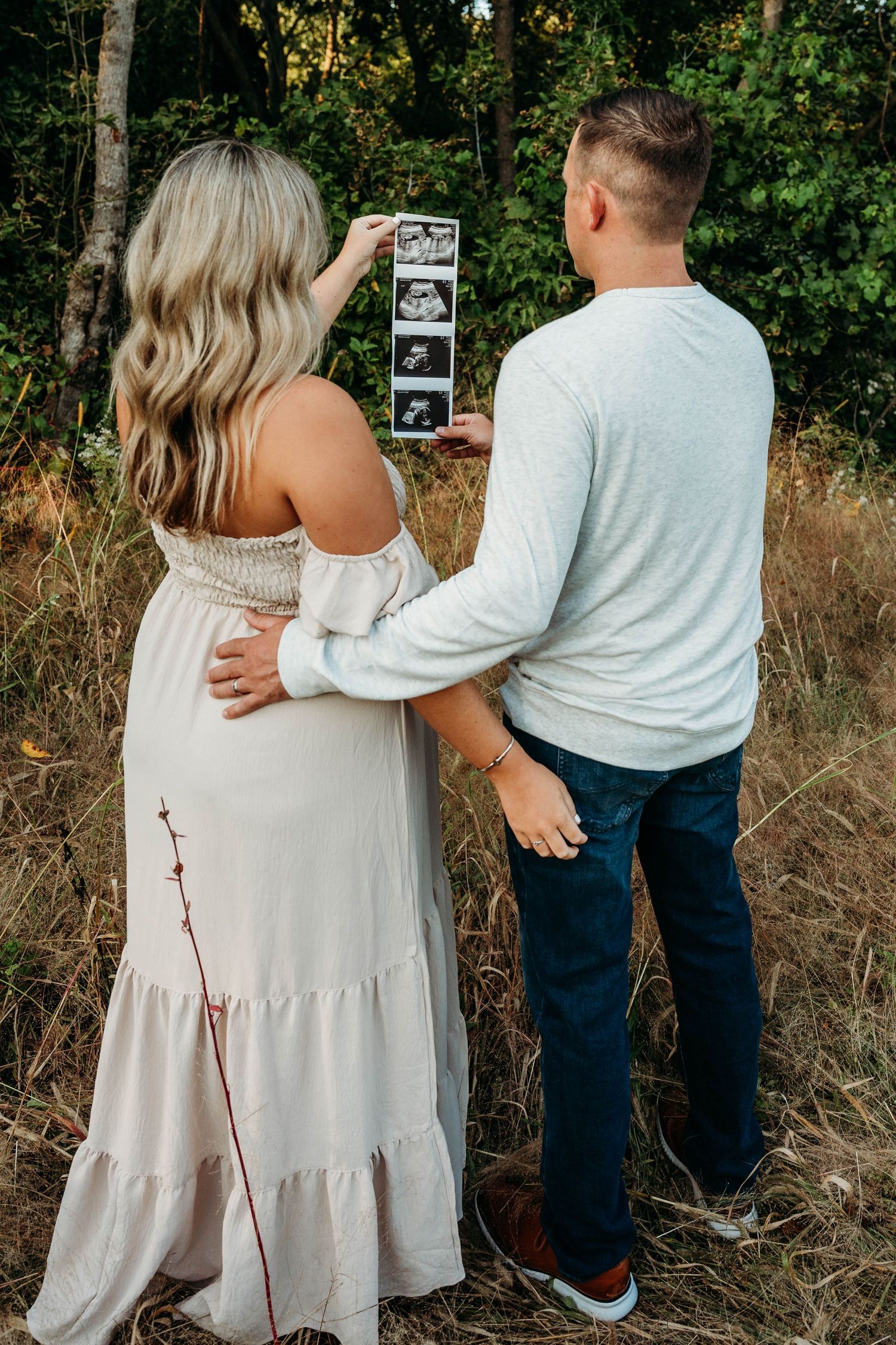 A couple in light colored clothing holds sonogram photos while standing together in a natural outdoor setting.