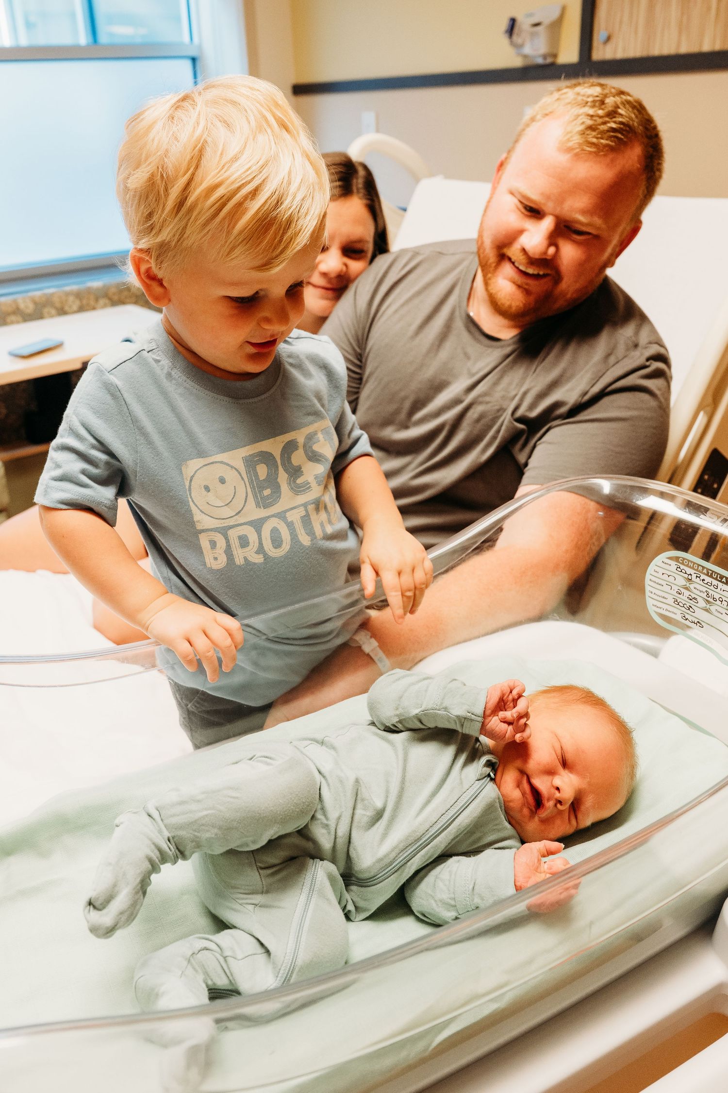 A toddler and adults lean over to interact with a baby lying on a changing table in a medical environment.