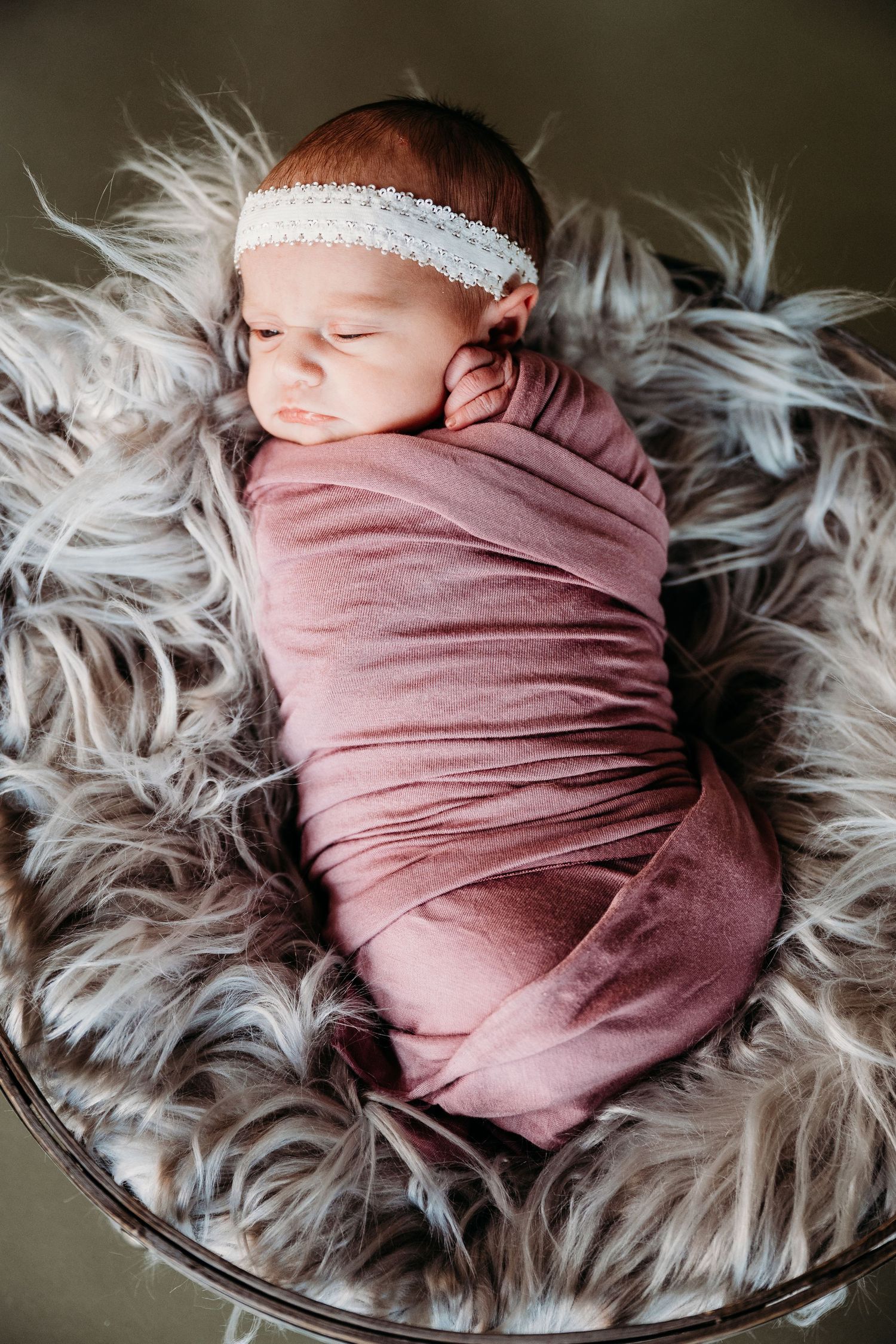 Sleeping newborn wrapped in dusty rose swaddle blanket rests peacefully on fluffy gray fur rug.
