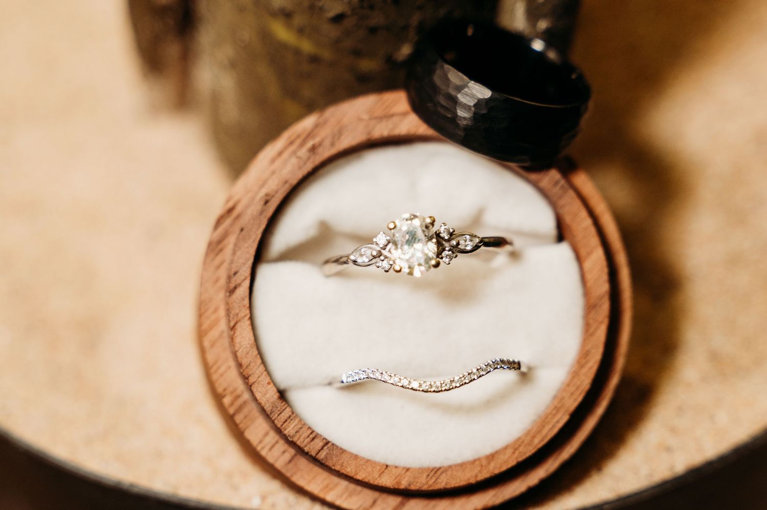 Close-up of a diamond engagement ring and curved wedding band nestled in a wooden ring box with white cushion.