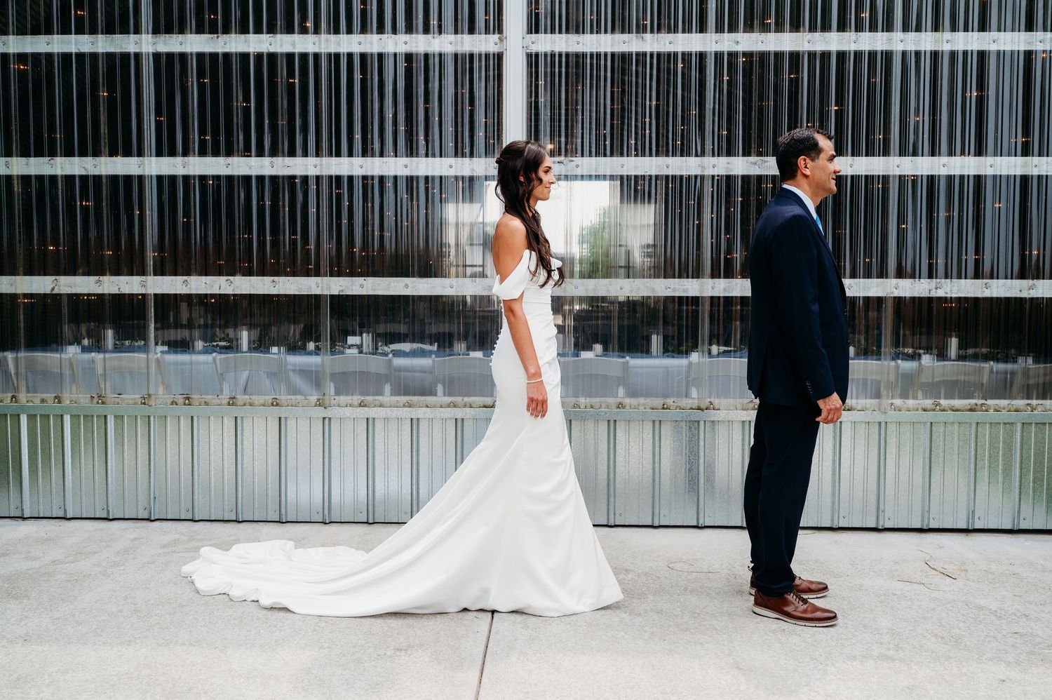 Bride in a flowing white gown and groom in navy suit share a first look moment outside a modern glass building.