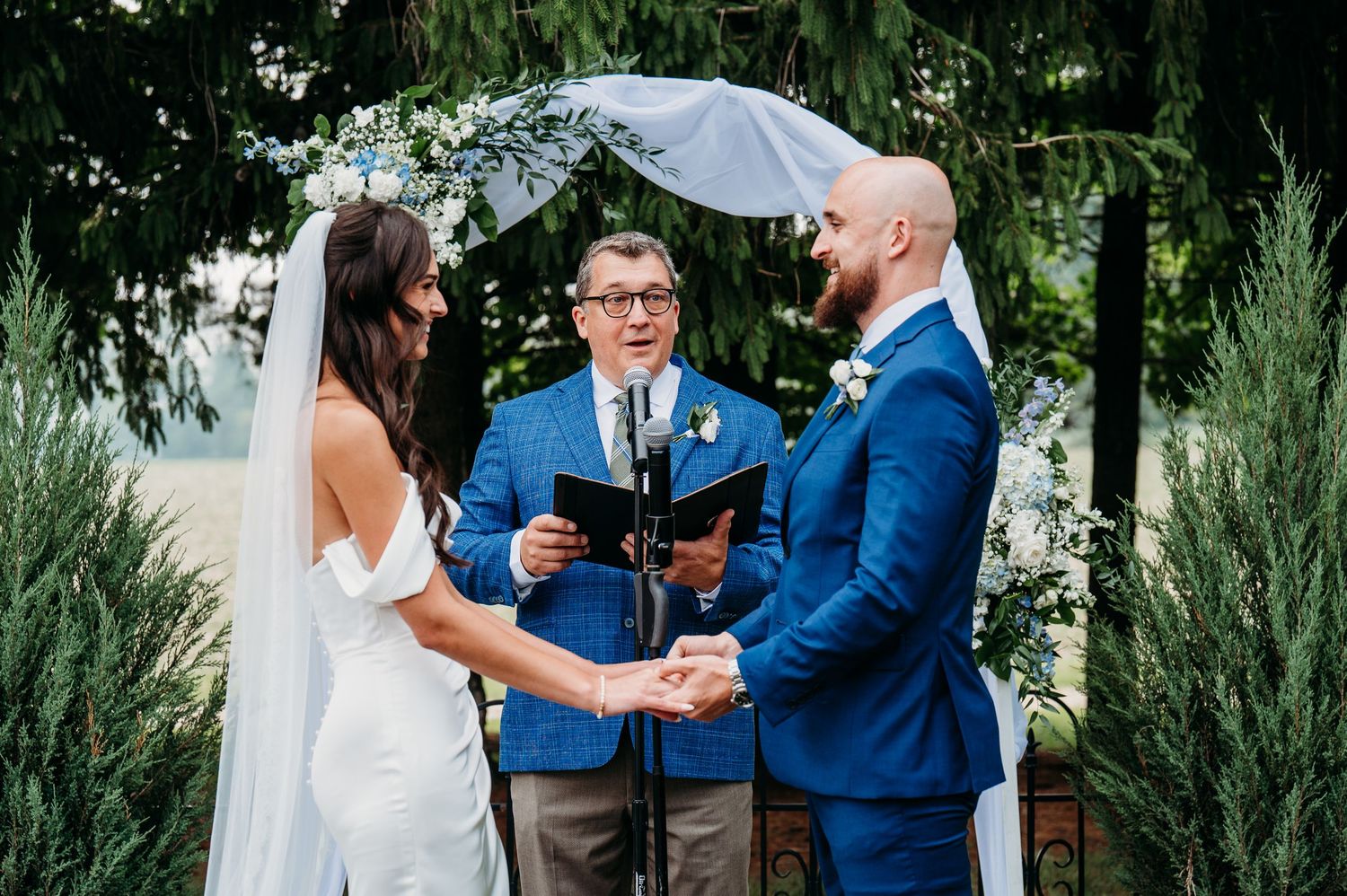 Couple exchanges vows outdoors with officiant in blue suit, bride in white gown, groom in navy suit, under floral arch.