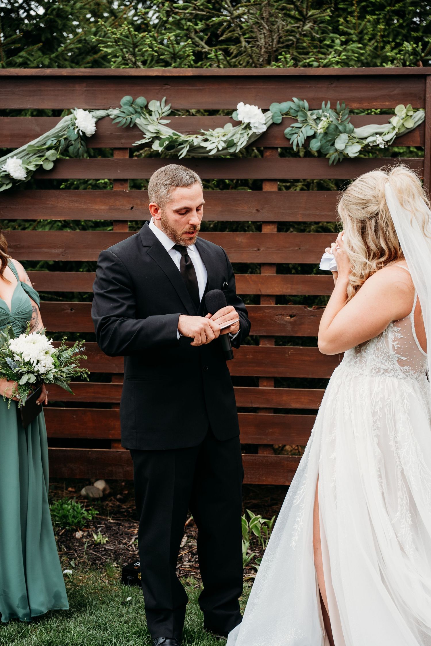 Groom places wedding ring on bride's finger during outdoor ceremony with rustic wood backdrop and greenery garland.