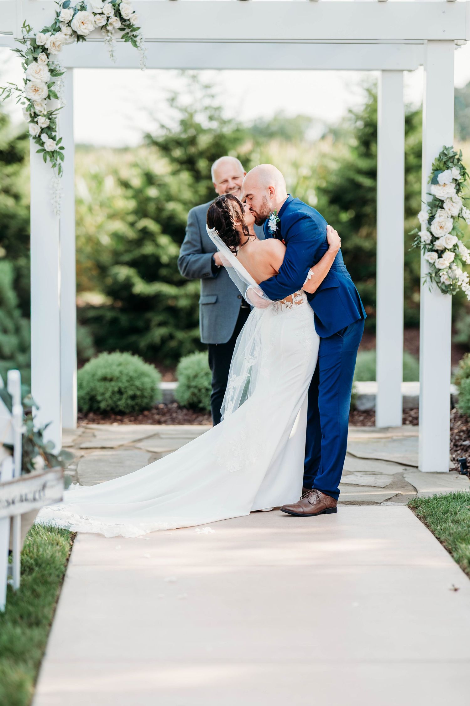 Bride and groom share first kiss under white floral arch at outdoor wedding ceremony with officiant smiling behind them. Blue Heron Barn