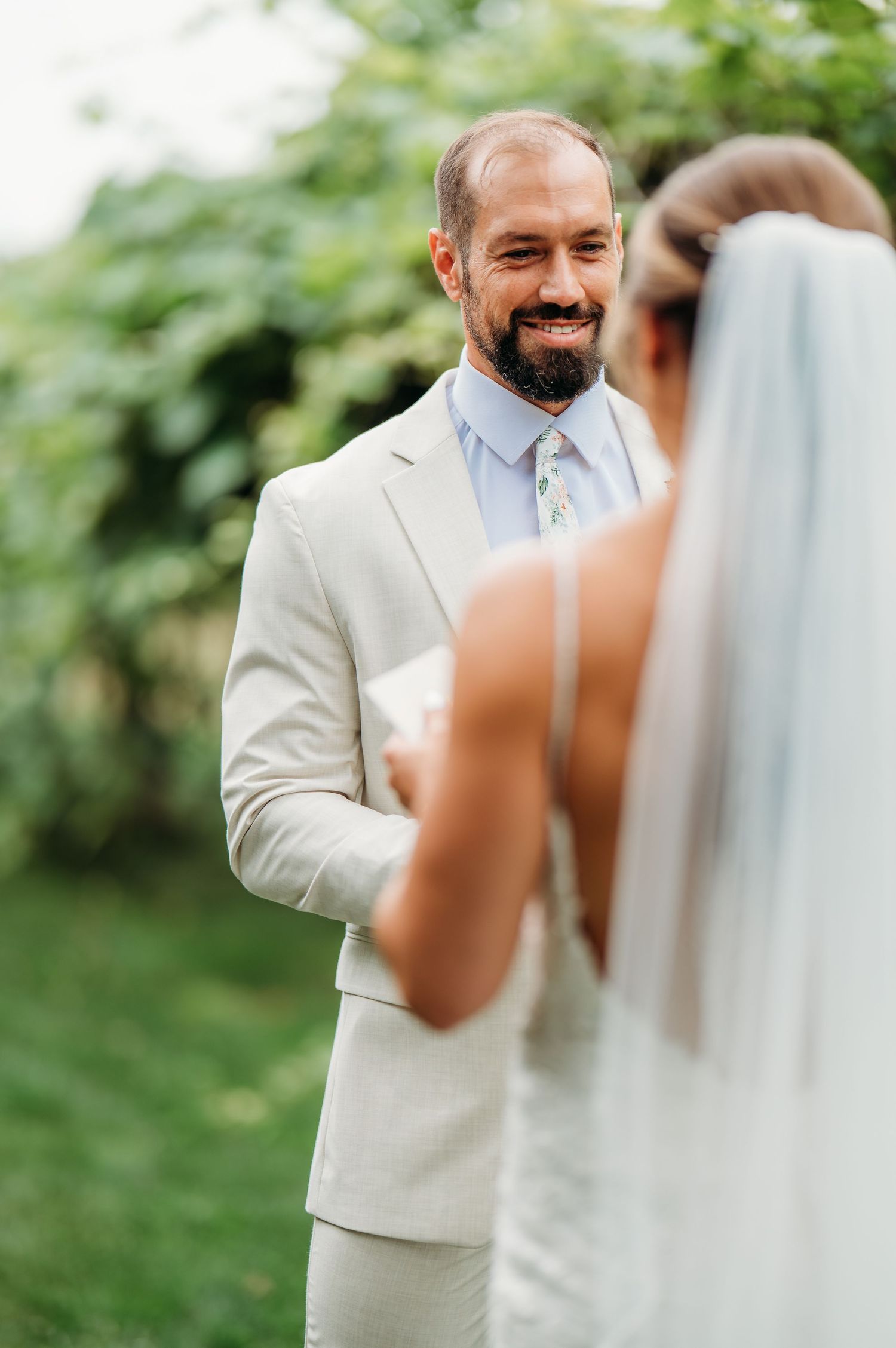 Groom in a light cream suit smiles emotionally during first look wedding moment in outdoor garden setting.