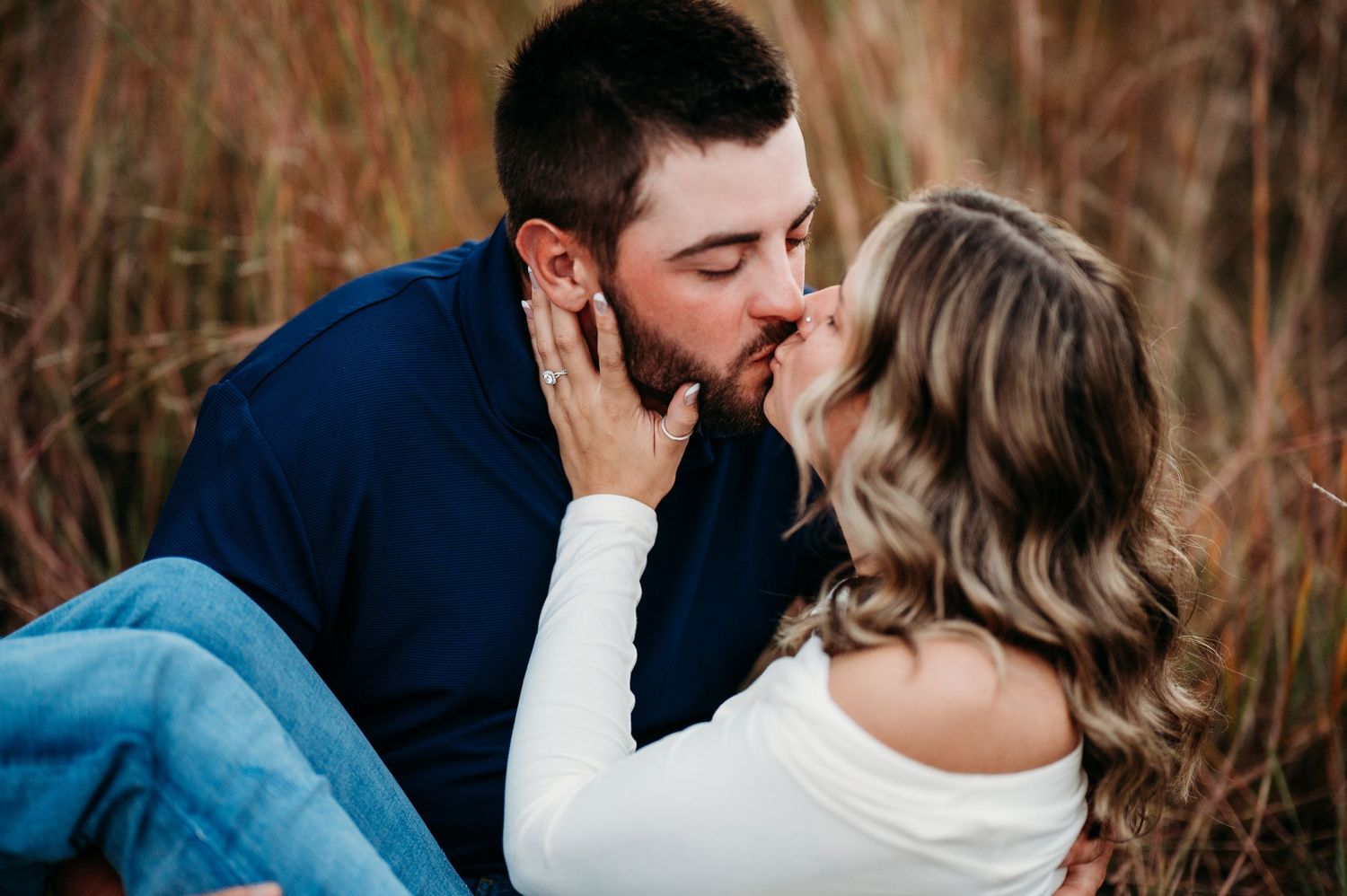 A romantic couple shares tender kisses and embraces in a field of tall grass during golden hour.