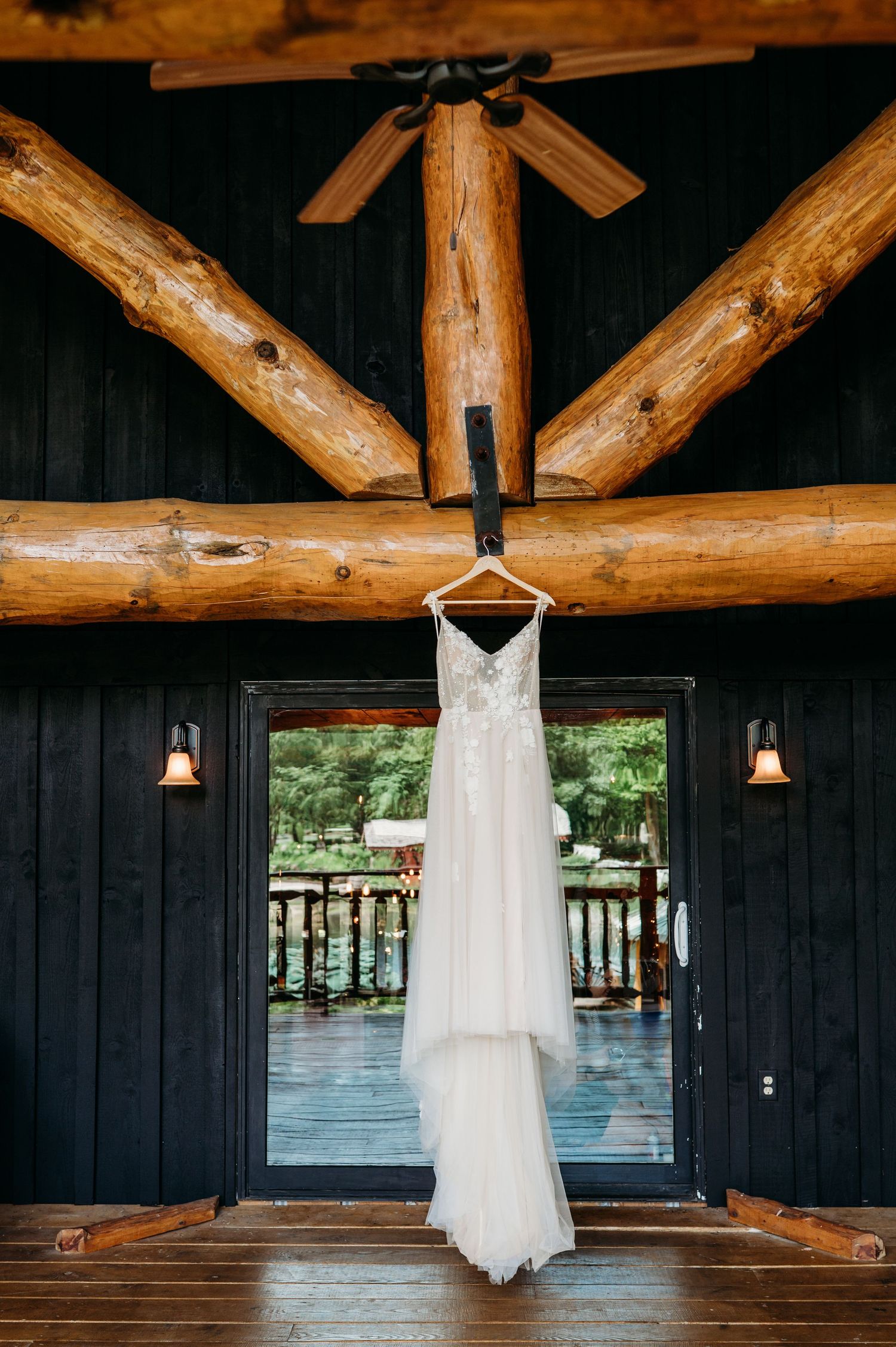 White wedding dress hanging from rustic wooden beams in a dark cabin-style venue with natural lighting.