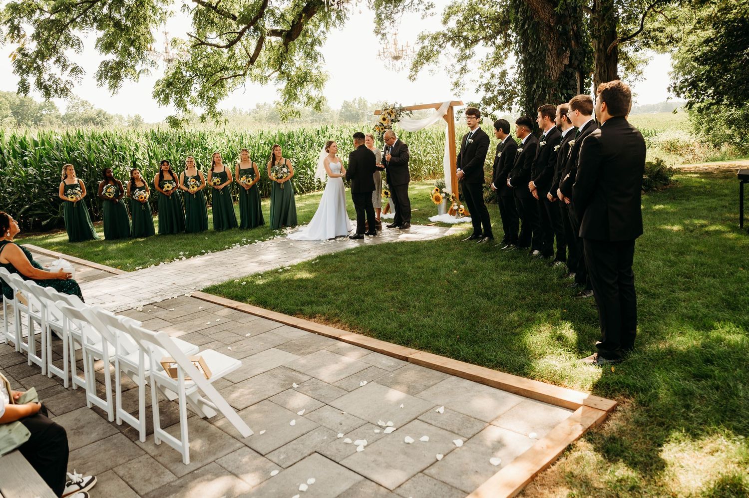 Outdoor wedding ceremony on stone patio with bridal party in green dresses and groomsmen in black suits under a large tree.