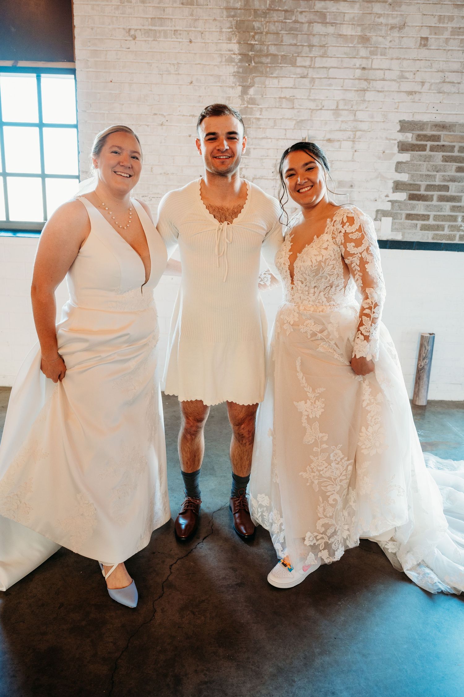 Wedding party members sitting together in white formal attire against an industrial brick wall background.