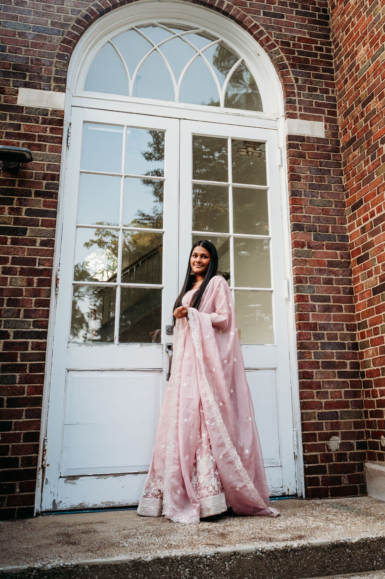 Traditional pink embroidered dress photographed against white arched doorway and red brick building exterior.
