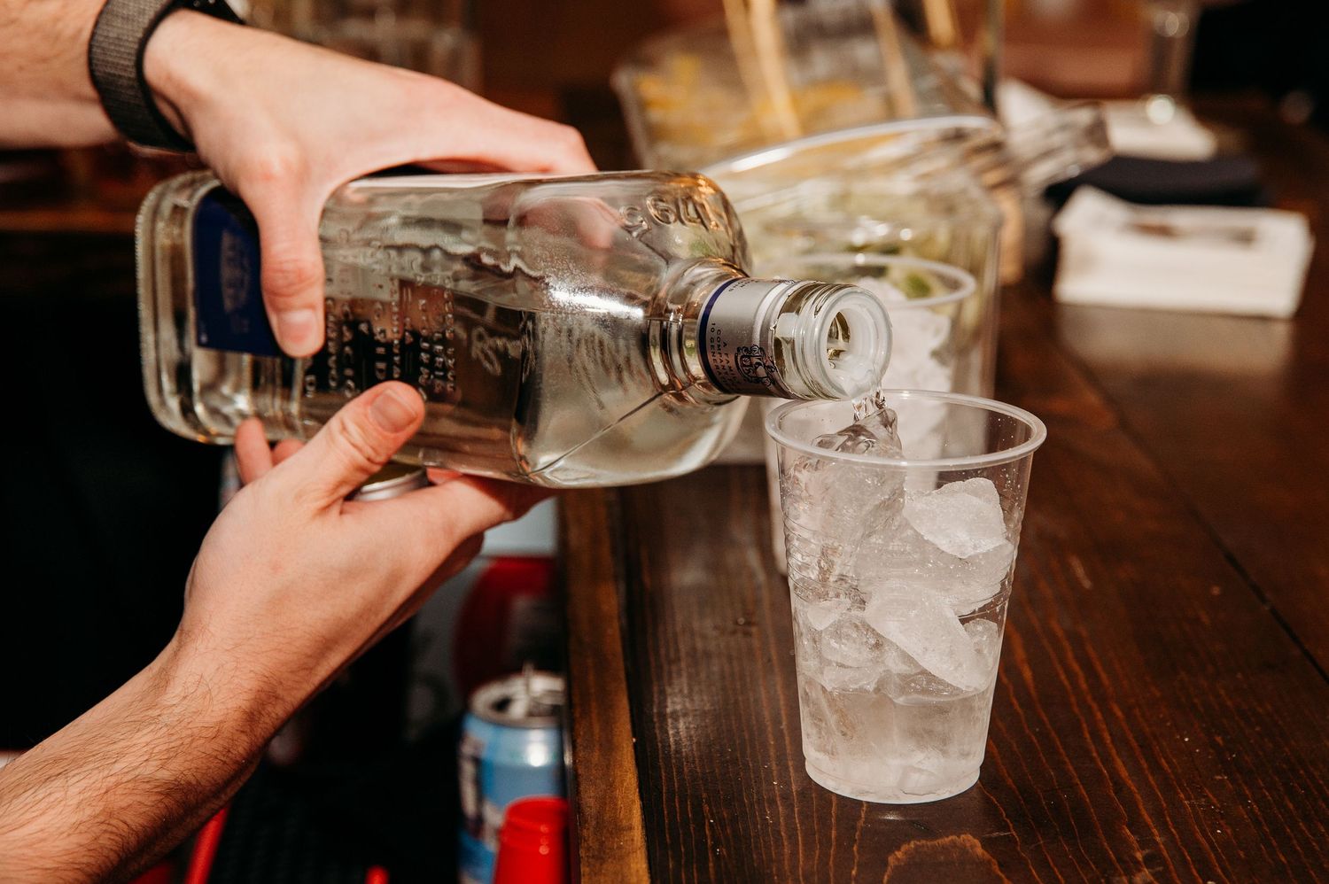 Someone pours clear liquor from a glass bottle into a tall glass filled with ice cubes on a wooden bar surface.