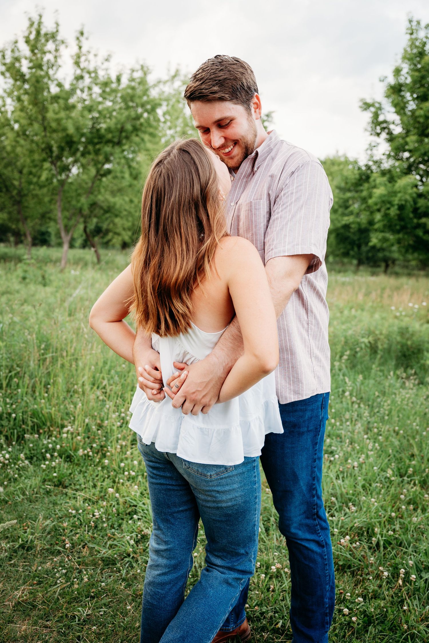 A couple embraces in a lush green field, the man smiling down at the woman in a tender outdoor moment.
