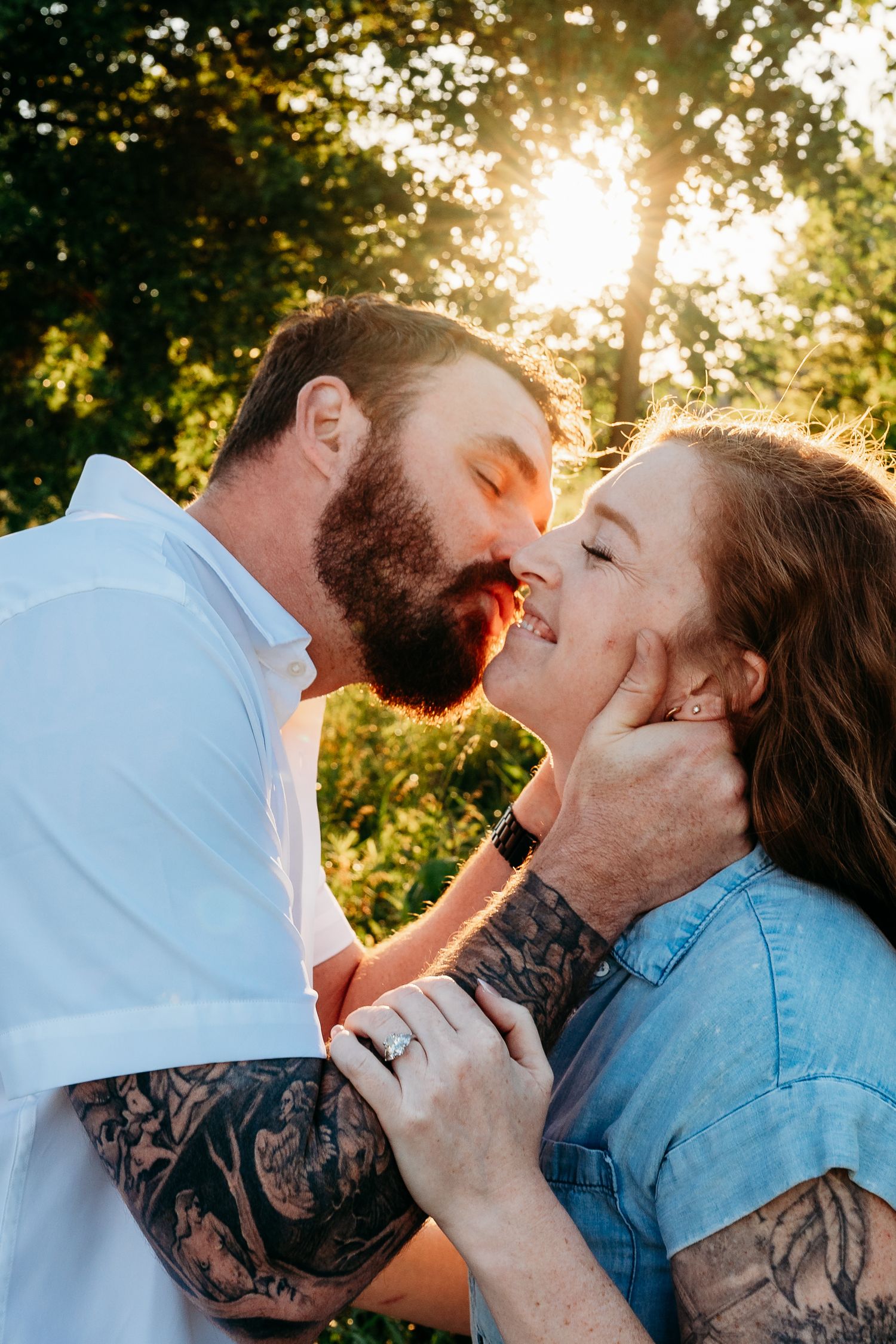 Tattooed couple sharing a romantic kiss outdoors at golden sunset, man tenderly holding woman's face in lush green park setting.