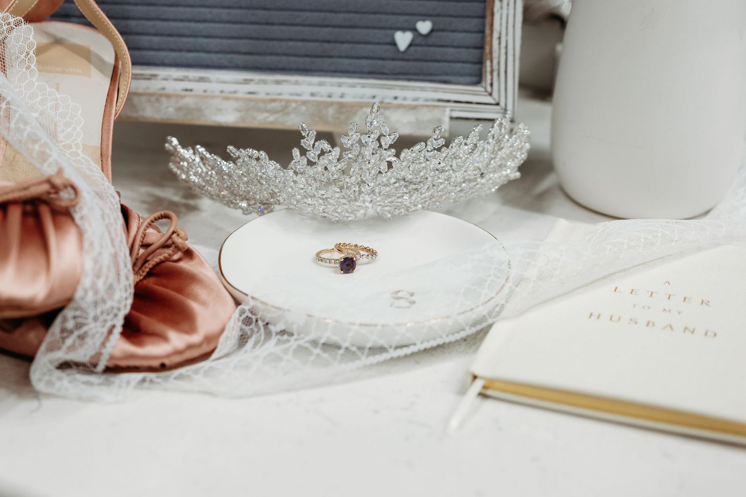 Elegant wedding flat lay with blush heels, crystal tiara, diamond ring on heart dish, veil, and a letters to husband journal.