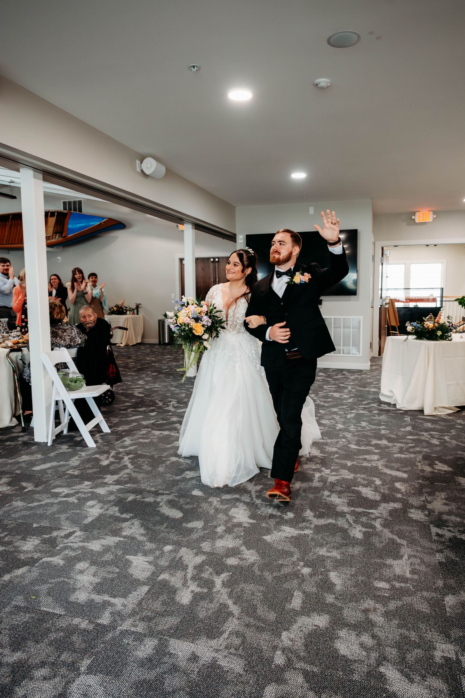 Bride and groom make grand entrance at wedding reception, groom waving to guests in decorated banquet hall.