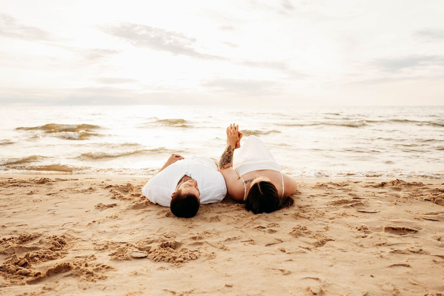 Romantic couple lying on sandy beach together, wearing white outfits with ocean waves in the background.