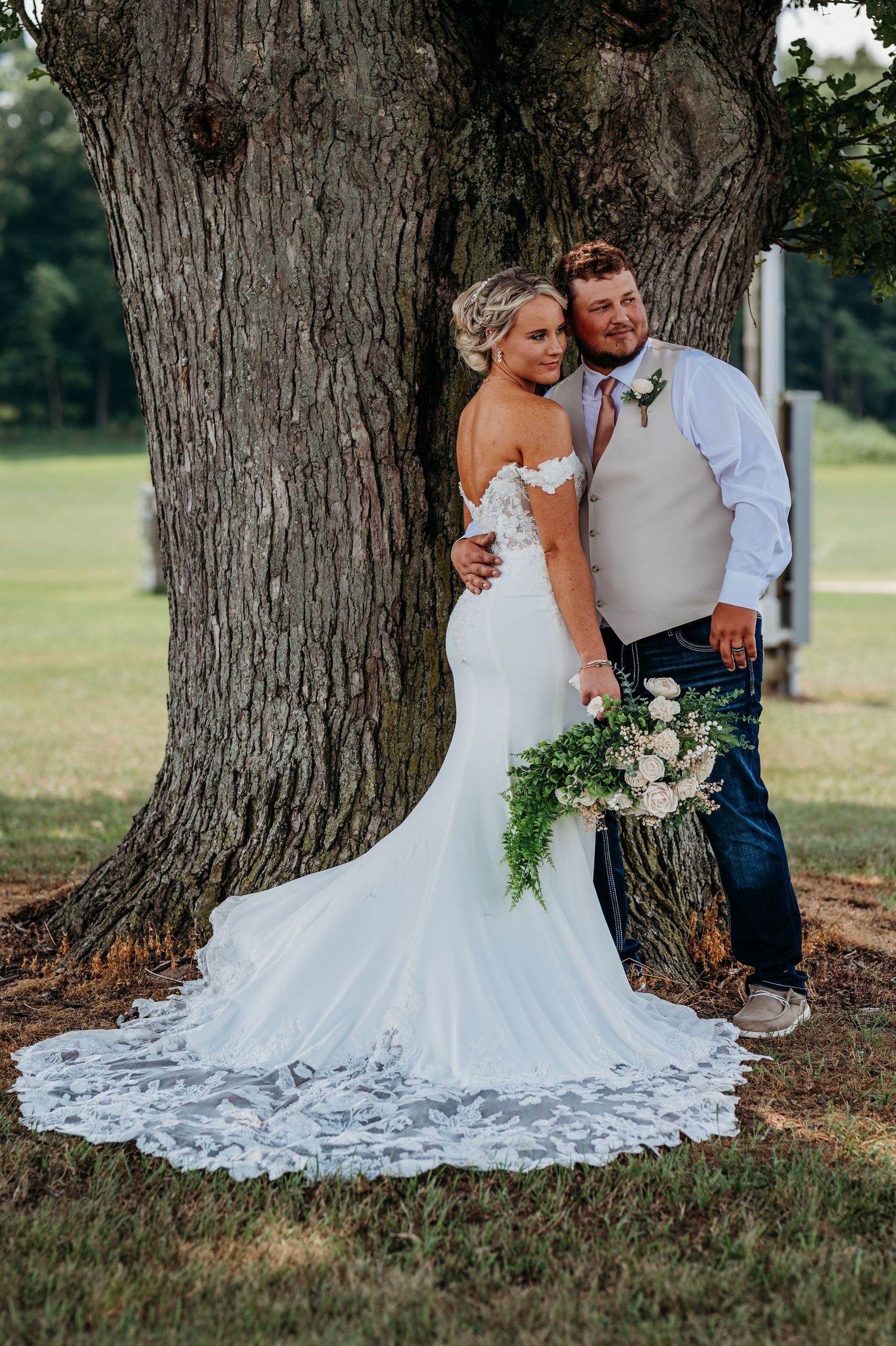 Newlyweds pose intimately against large tree trunk with bride's lace dress spread beautifully on grass.
