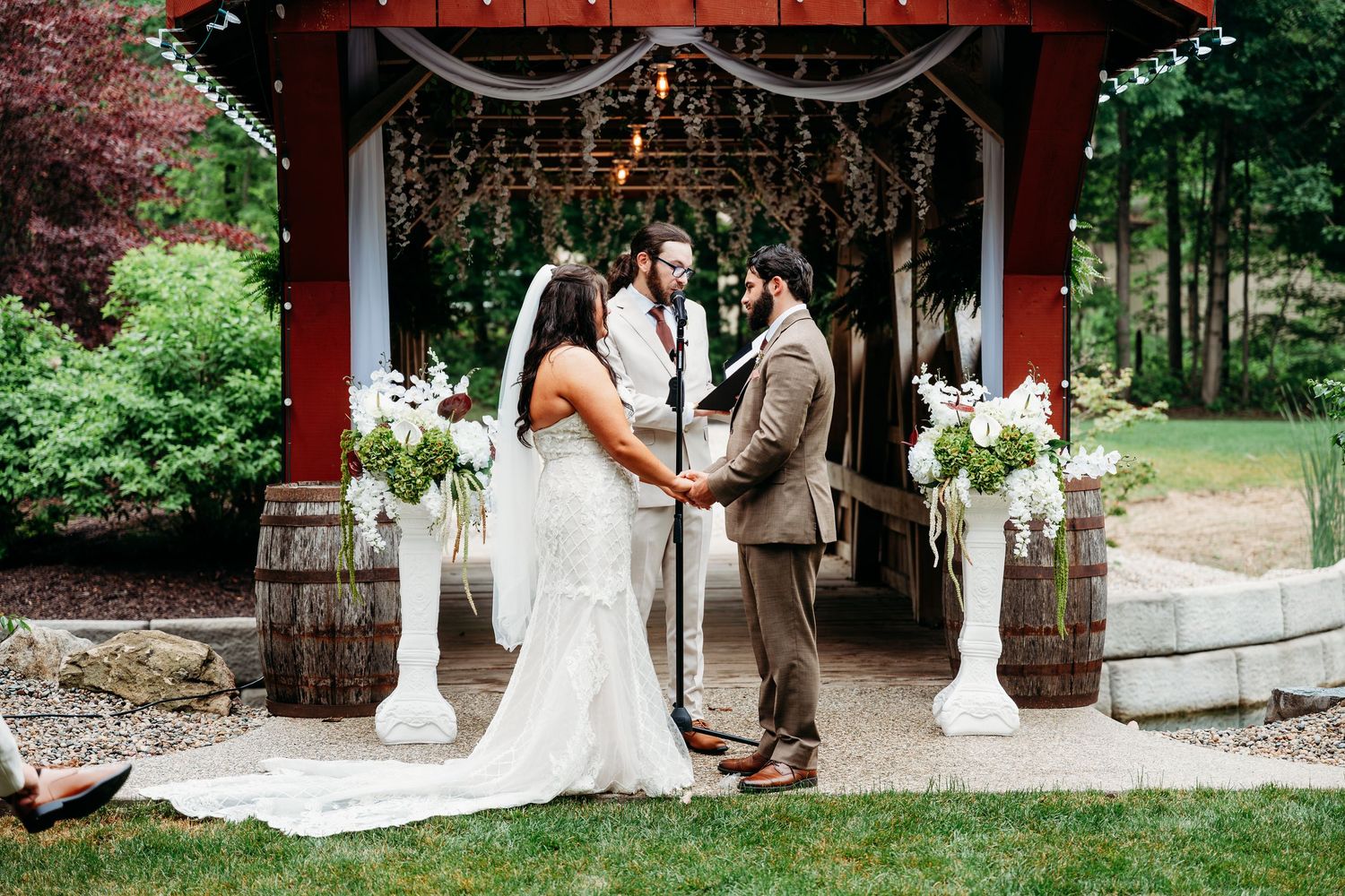 Couple exchanges vows at rustic outdoor wedding ceremony with red barn backdrop and floral arrangements.