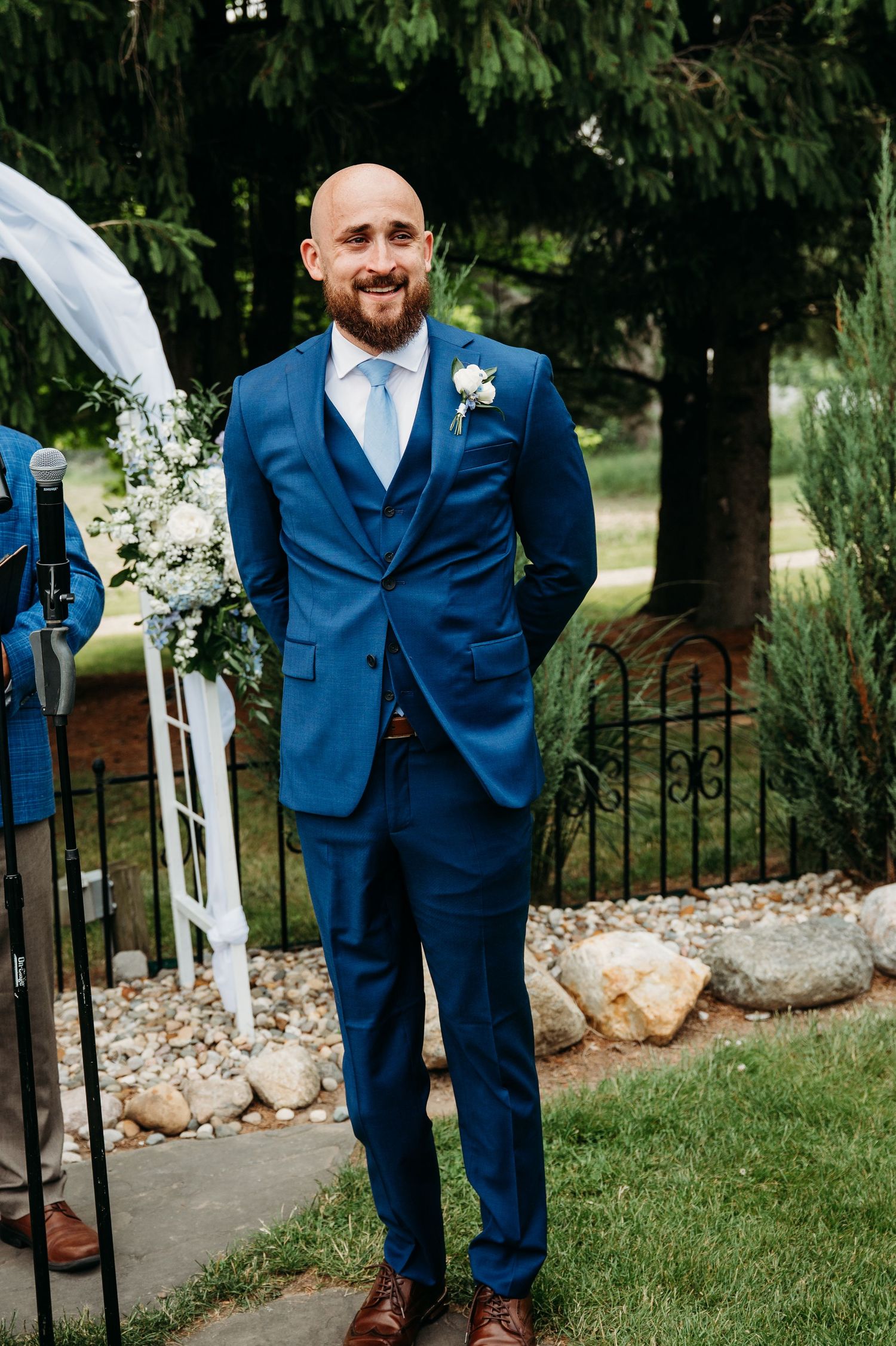 A groom in a navy blue suit poses playfully on a garden path with decorative stone landscaping and greenery.