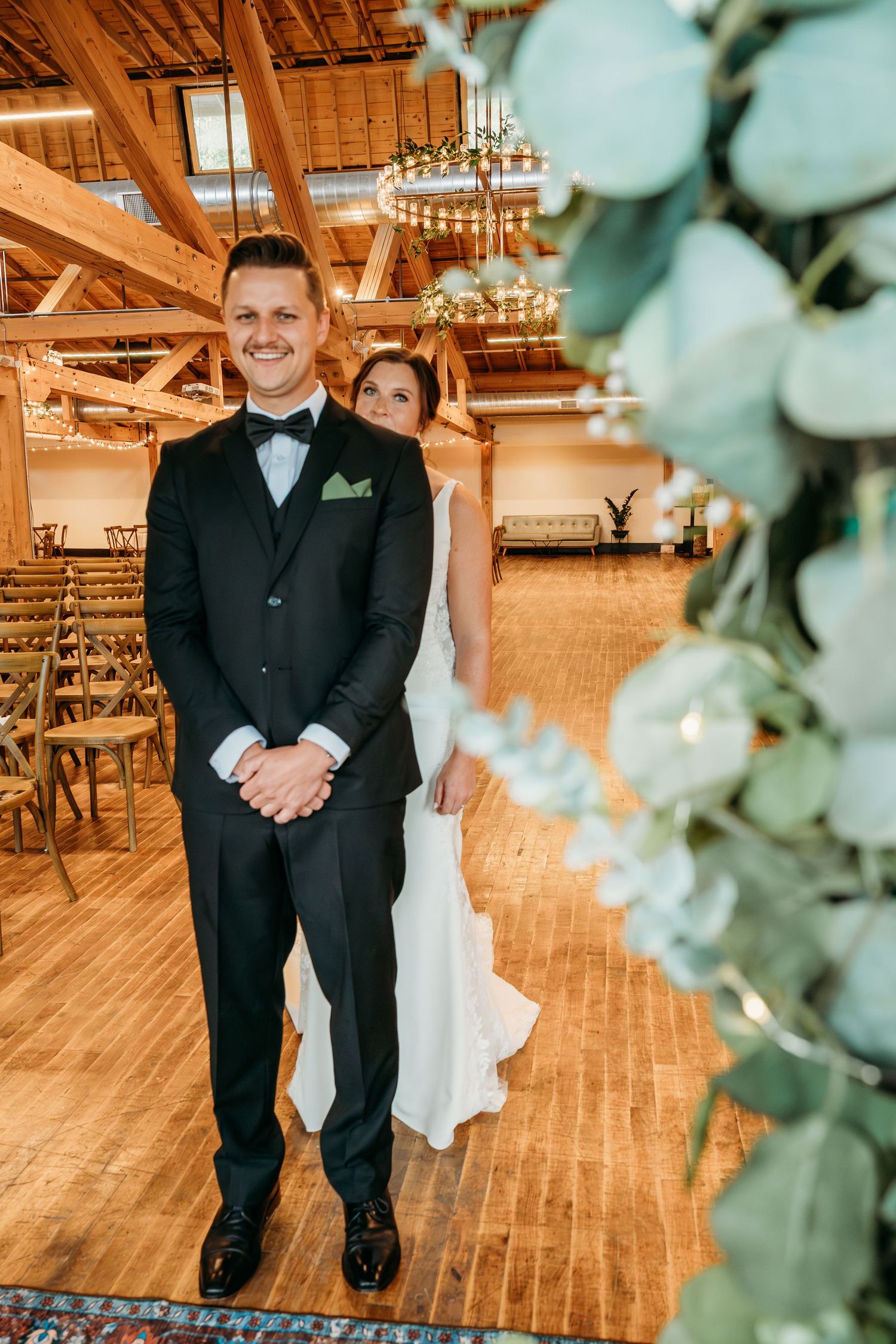 Wedding couple stands on rustic wooden floor in elegant venue with exposed beams and romantic lighting.