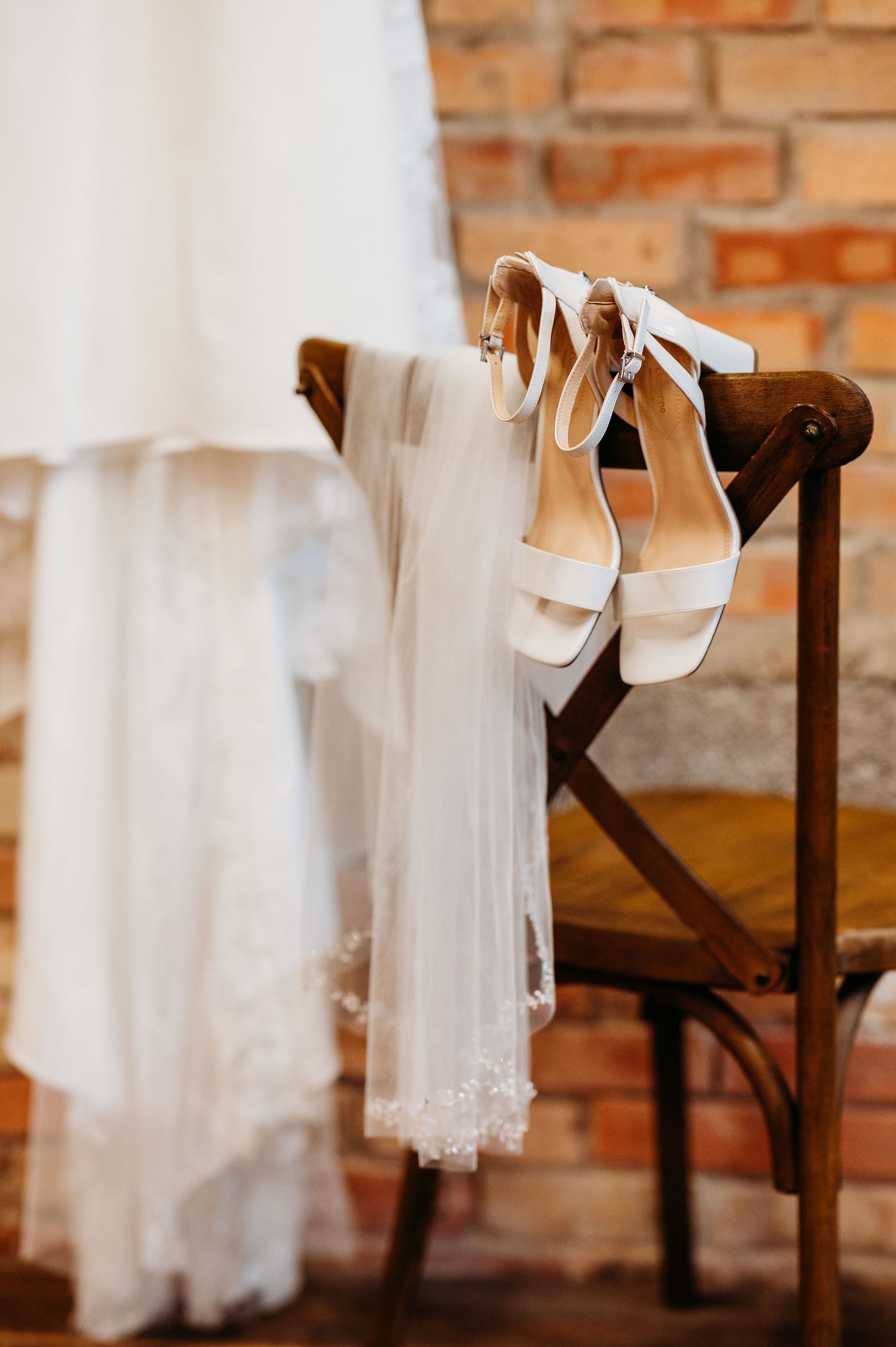 White wedding shoes rest elegantly on a wooden bench against a rustic brick wall.