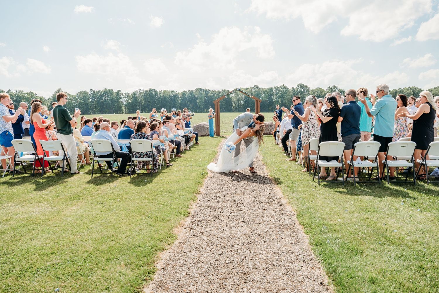 Outdoor wedding ceremony on green lawn with guests seated in white chairs along gravel aisle, couple kissing near waterfront.