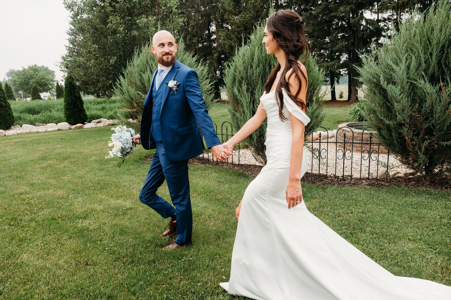 A bride and groom hold hands while walking through a lush green garden, she in a white gown, he in a blue suit.