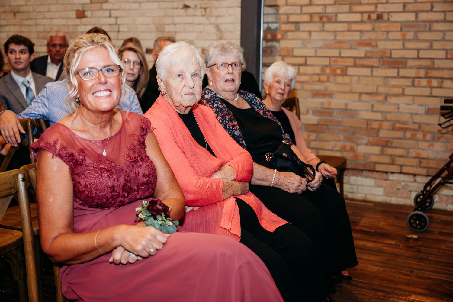 Elderly women smiling and watching a wedding ceremony while seated in wooden chairs inside a brick venue.