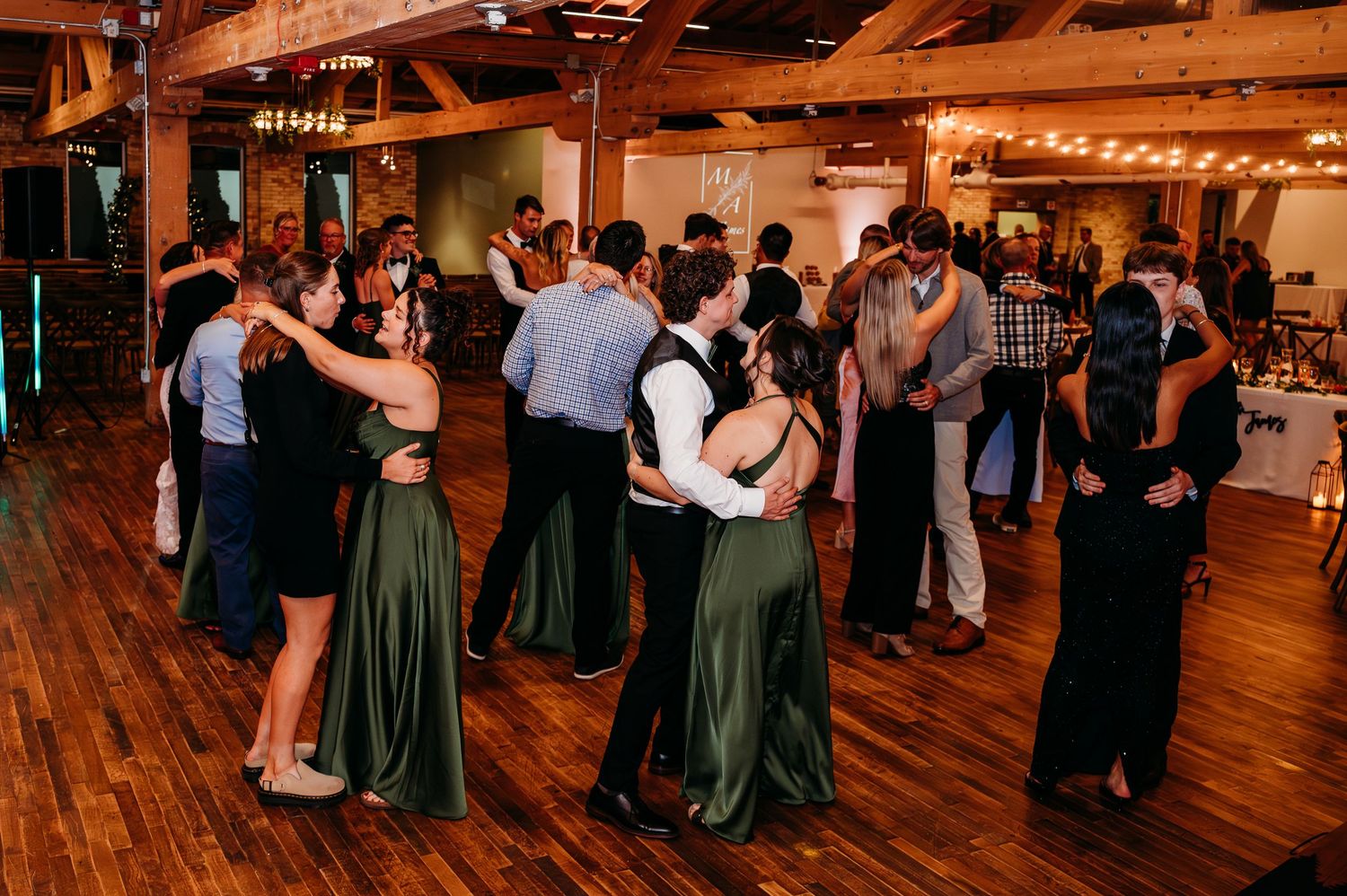 Couples dancing together on a wooden dance floor at a rustic wedding reception with warm string lights.