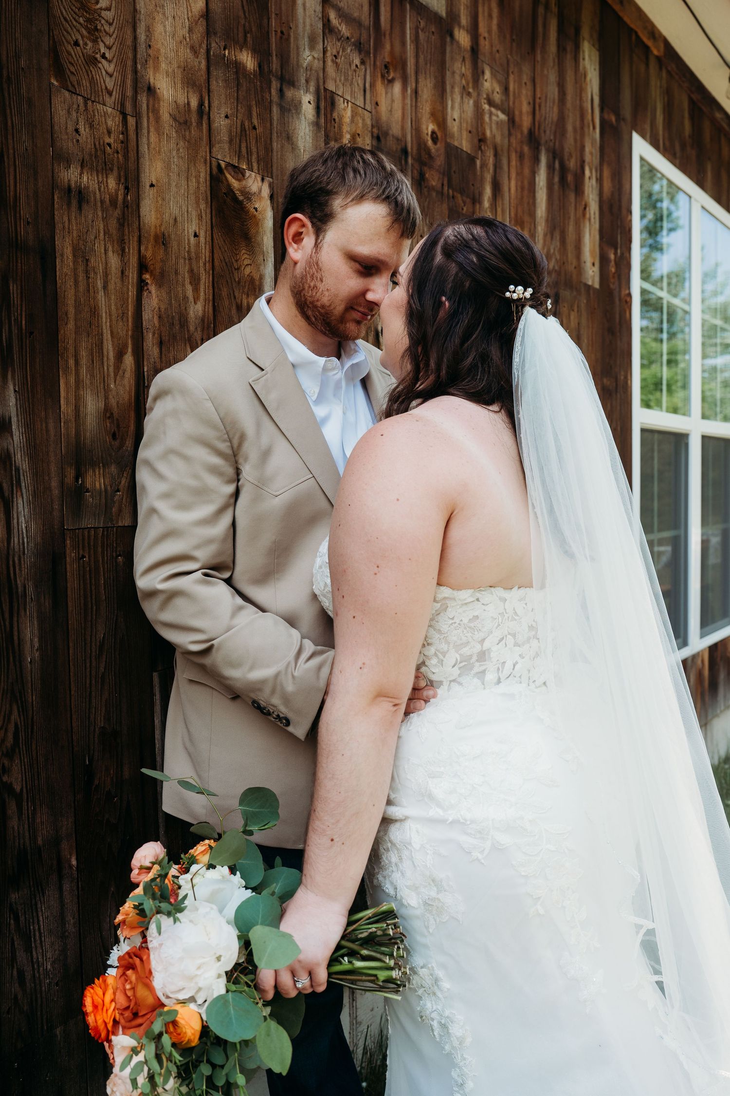 A tender wedding moment between newlyweds by a rustic wooden barn wall as the bride holds a vibrant orange and white bouquet.