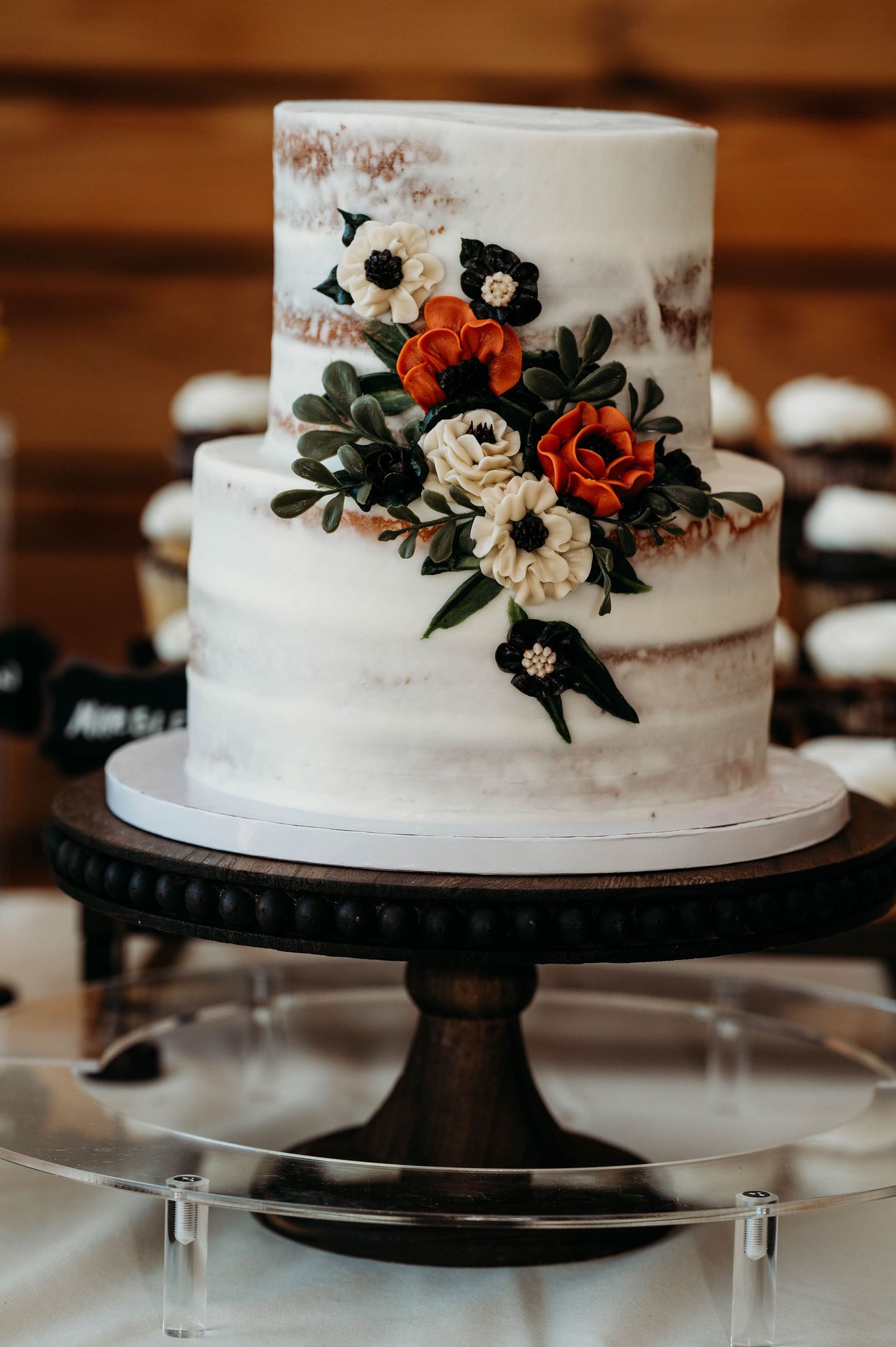 A rustic white wedding cake decorated with red and white sugar flowers on a dark pedestal stand.