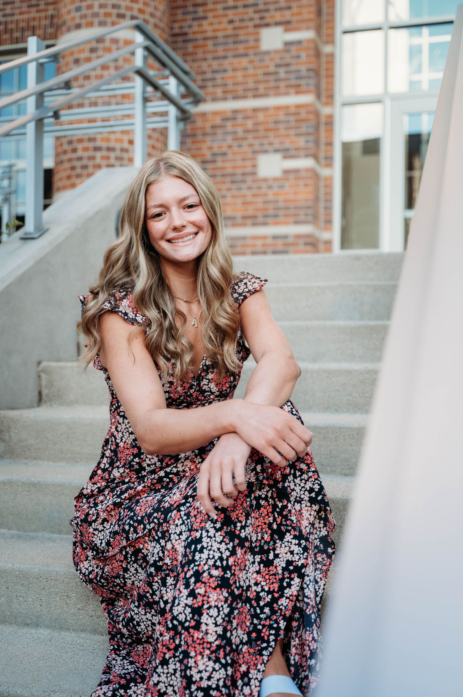 Smiling figure in a floral maxi dress sits casually on concrete steps outside a brick building.