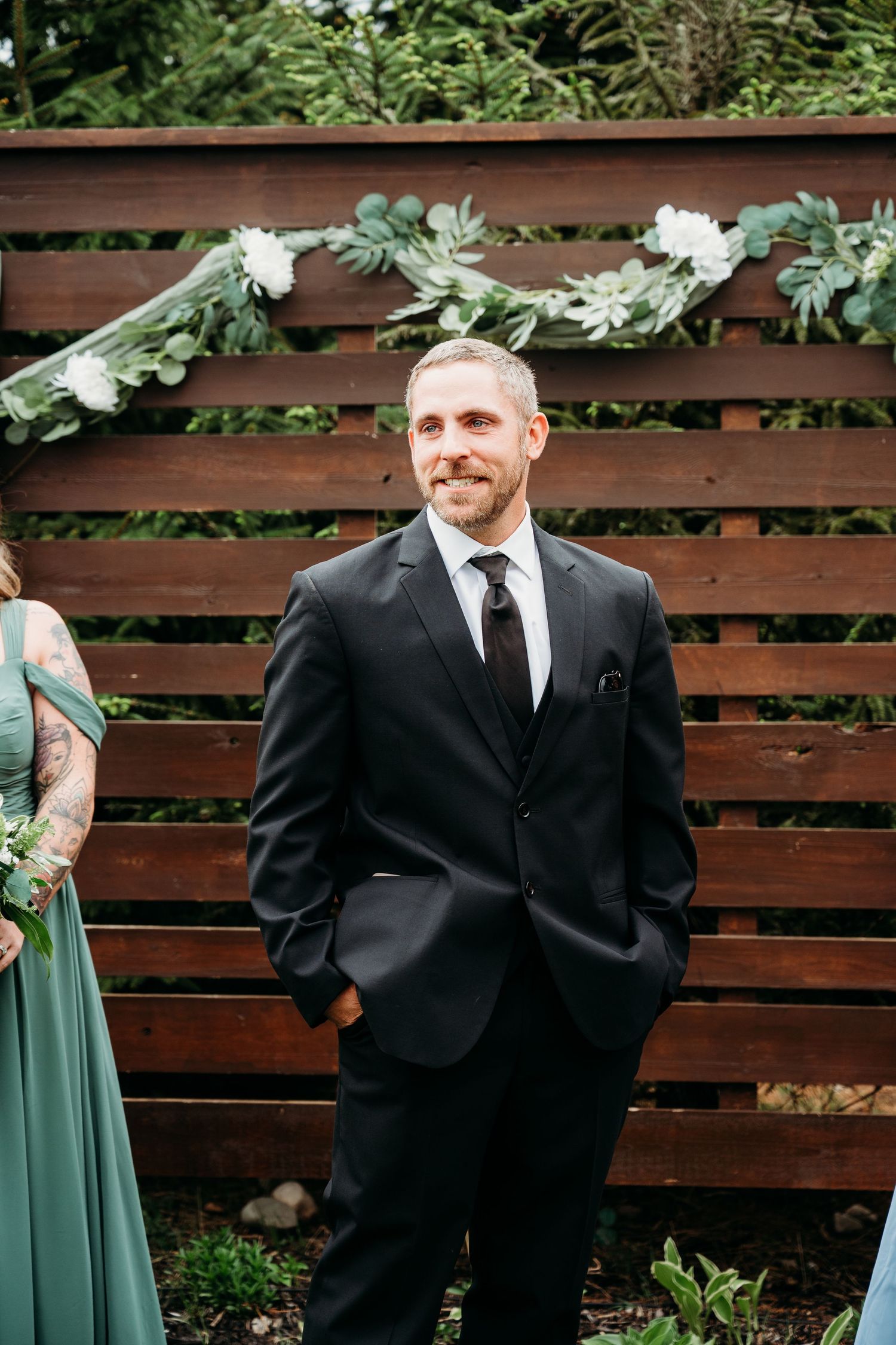 Groom in black suit and tie smiling at outdoor wedding ceremony with wood backdrop and white floral garland.