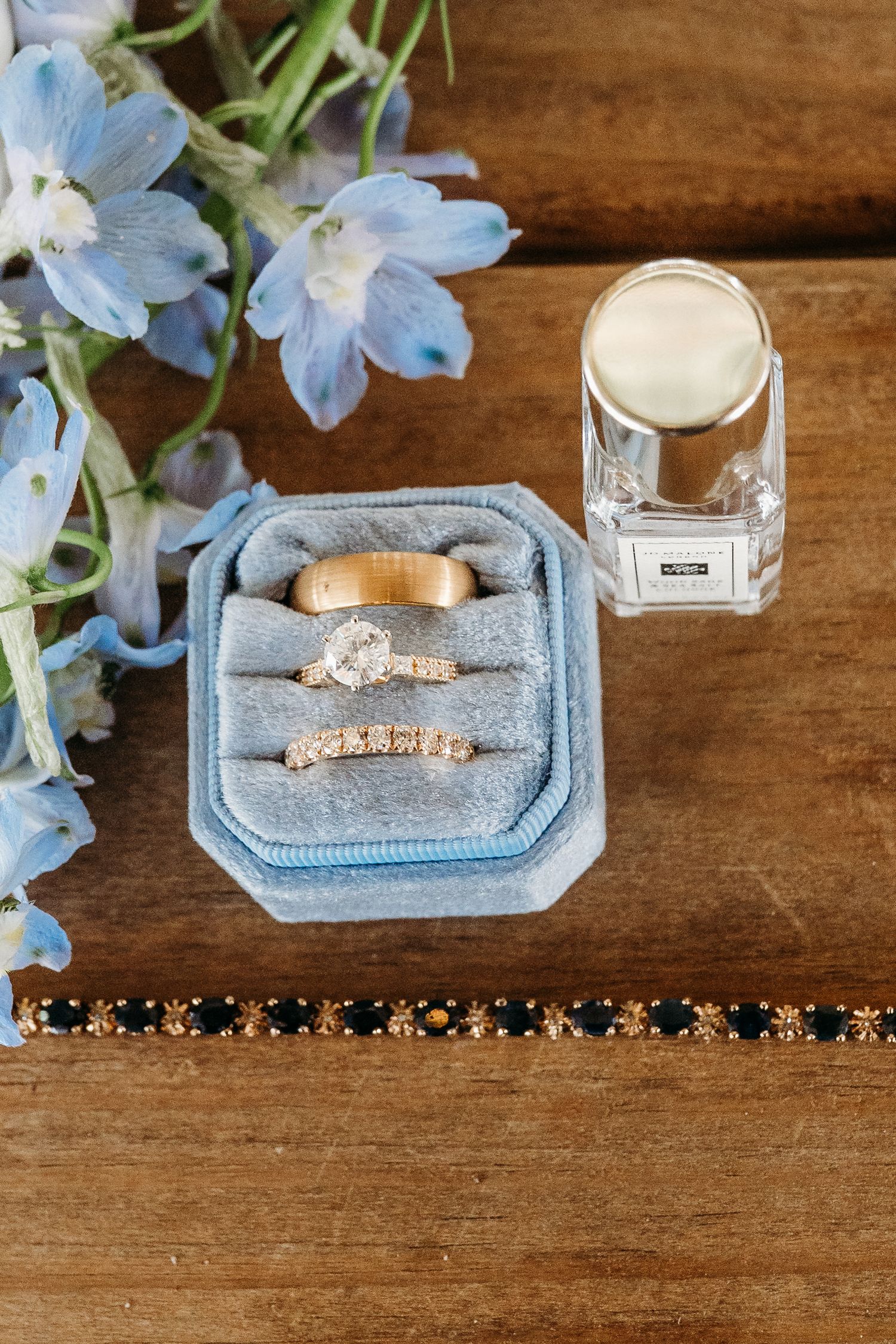 Blue velvet ring box with gold wedding bands and diamond engagement ring, beside perfume bottle and blue flowers on wood.