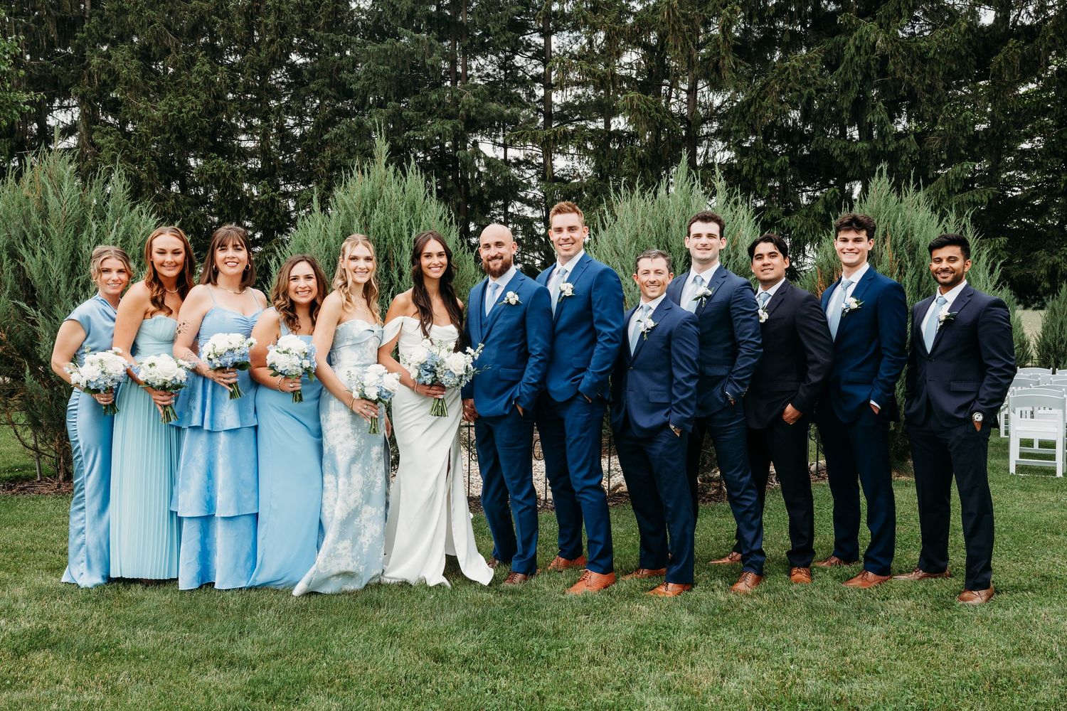Wedding party posing outdoors, bridesmaids in light blue gowns and groomsmen in navy suits flanking bride and groom.