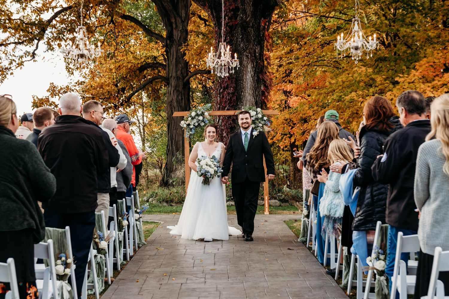 Bride and groom walk down outdoor aisle surrounded by fall foliage and chandelier decor at autumn wedding ceremony.