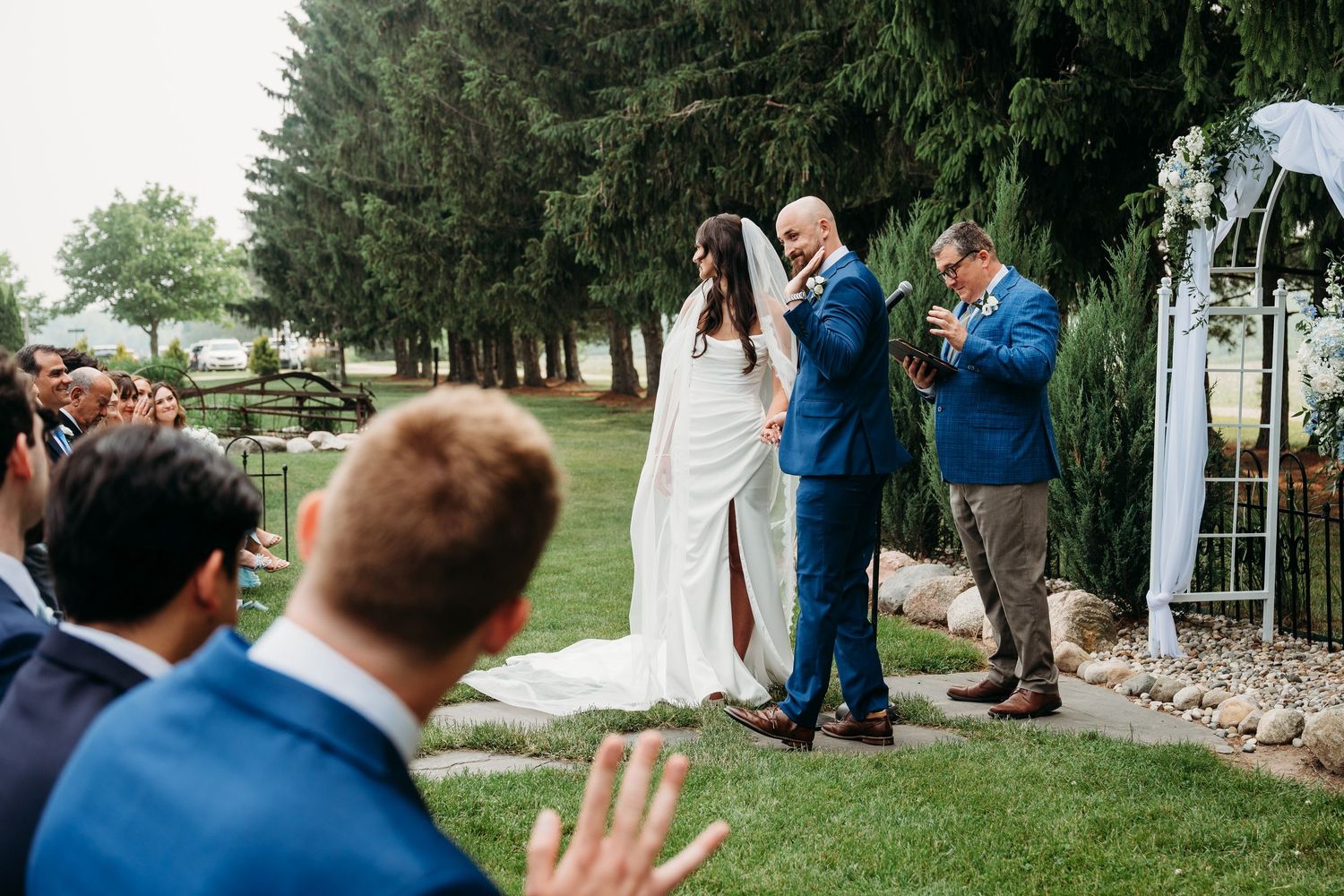 People gather for an outdoor wedding ceremony under a white arch surrounded by evergreen trees on a grassy lawn.