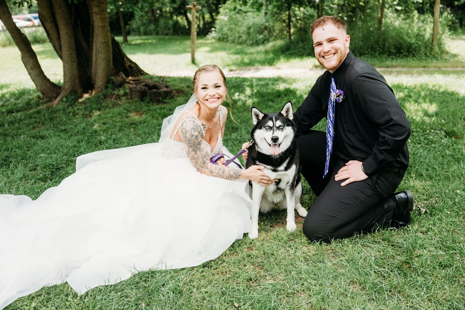 A couple in wedding attire kneels on grass with their Husky dog during outdoor wedding photography.