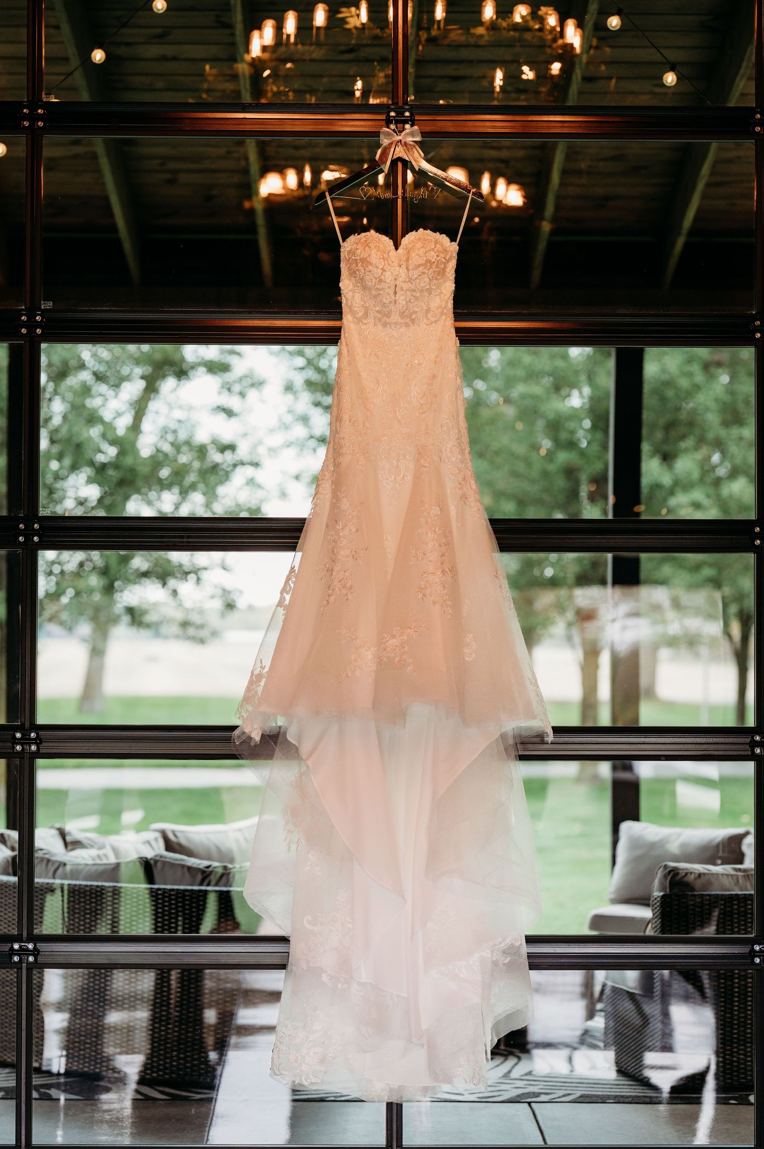 White wedding dress hanging in a rustic venue with string lights and large windows overlooking green outdoor scenery.