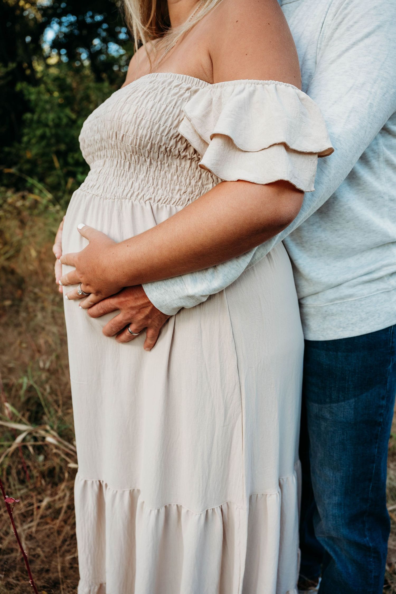 A close-up shot of a pregnant belly in a white off-shoulder dress being embraced from behind in an outdoor setting.
