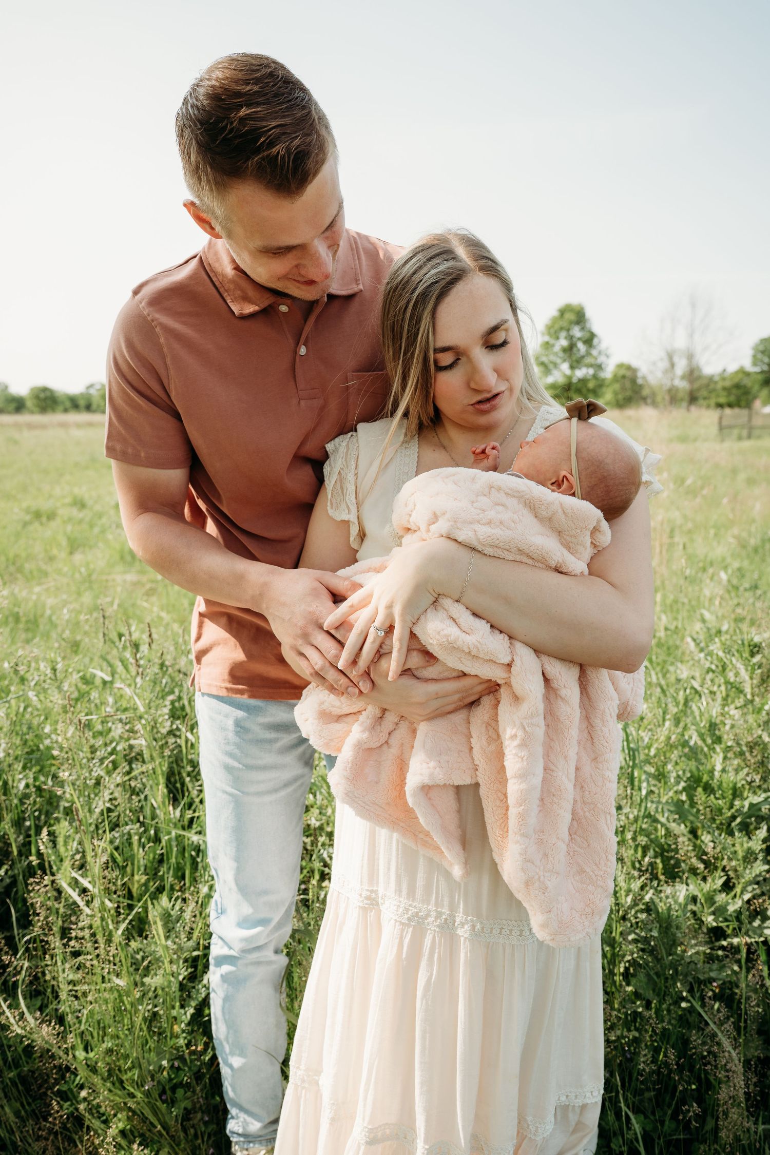 A couple embraces tenderly in a sunlit meadow wearing soft pink and cream colored outfits.
