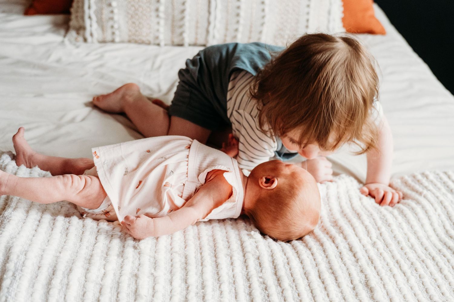 Tender sibling moment captured on white bedding with baby in pink dress.