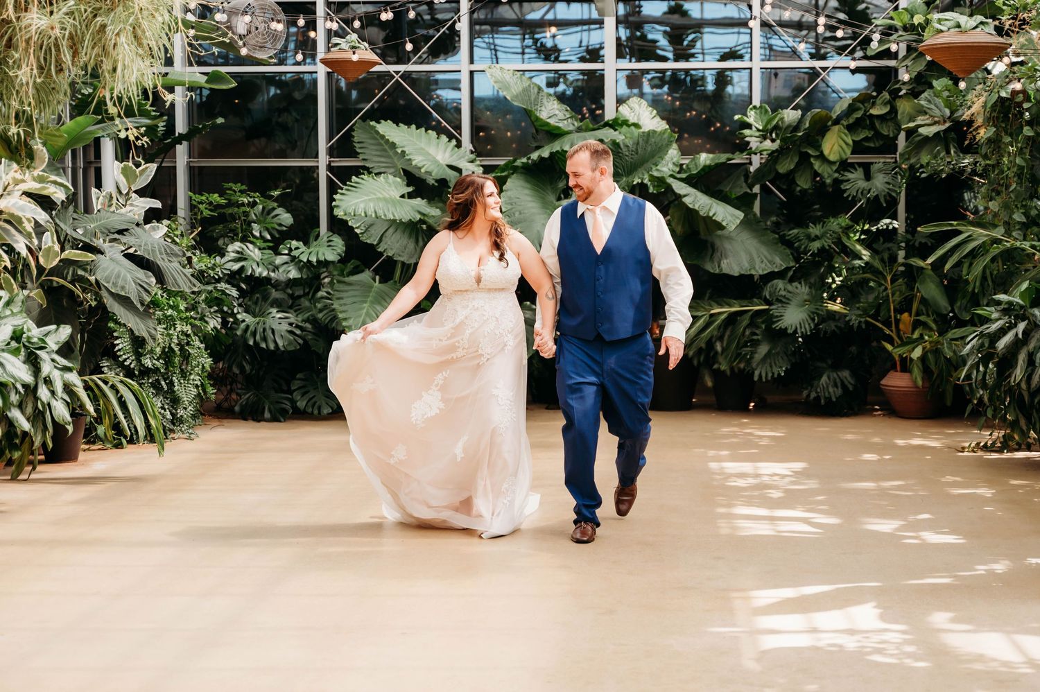 Two people walking together in a lush greenhouse setting with tropical plants and natural lighting.