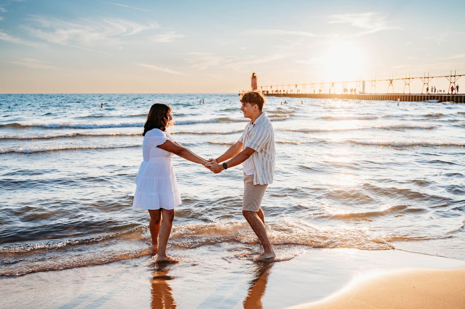 A couple holds hands while standing in the waves at sunset on a beach with a pier in the background.
