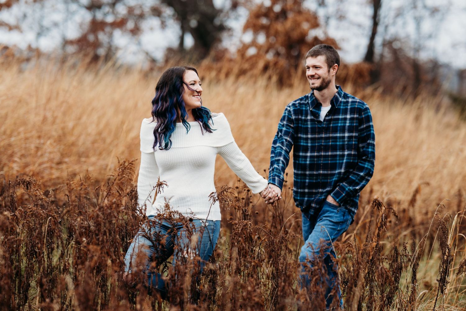 A couple walks hand in hand through a golden autumn field while sharing a candid laugh together.