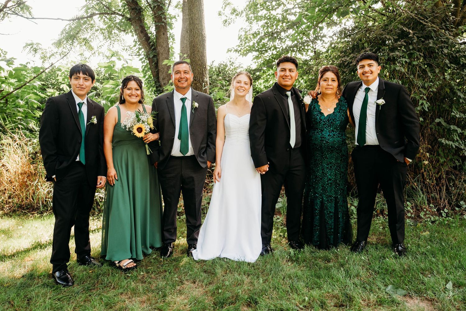 Wedding party poses outdoors in green and black formal attire with bride in white gown holding bouquet.