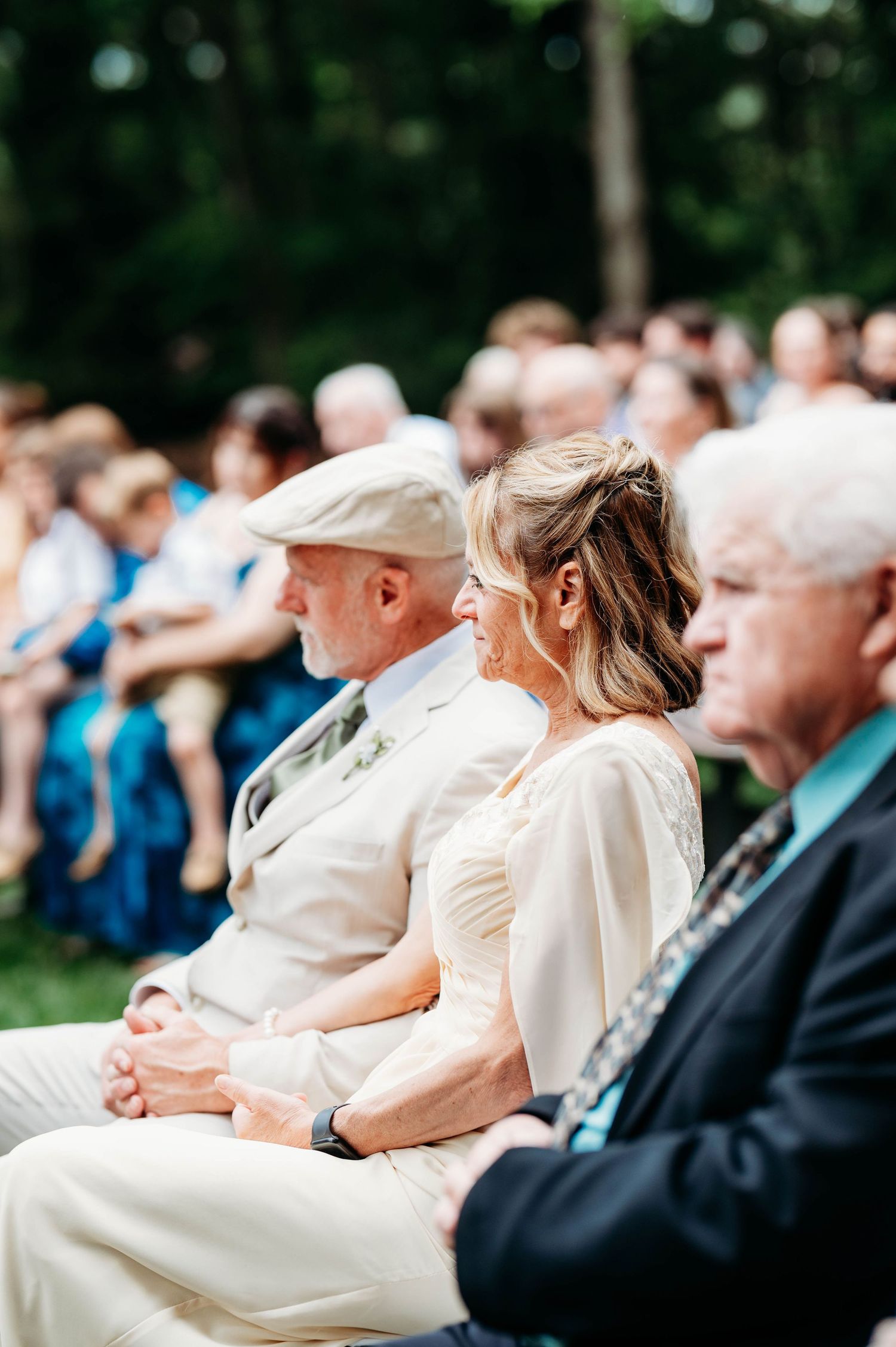 Wedding guests seated outdoors smiling during a garden ceremony, with bridesmaids in blue dresses visible in the background.