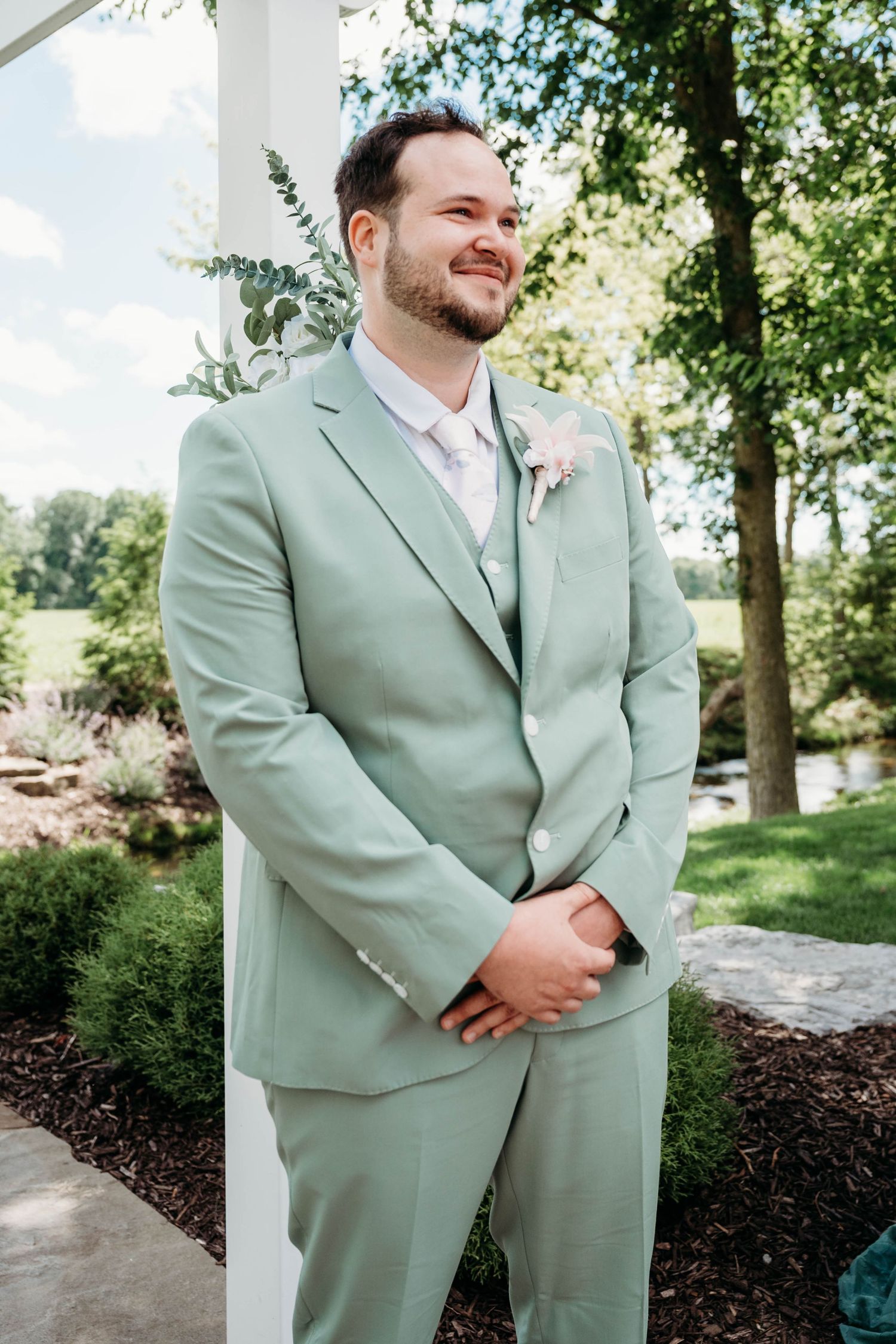 Smiling groom in sage green suit with boutonniere standing outdoors at wedding venue surrounded by lush greenery.