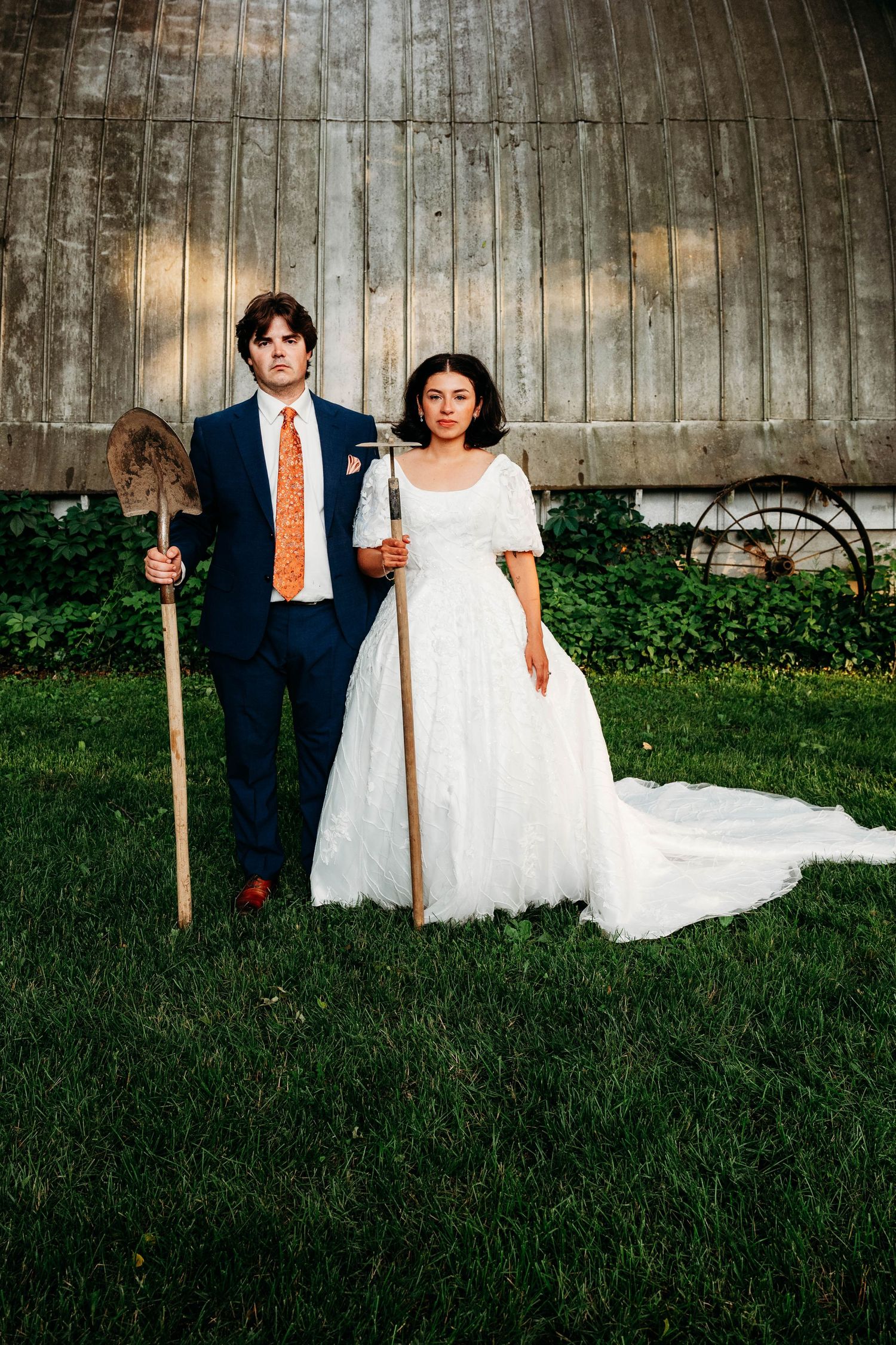 Bride and groom pose seriously holding farm tools before rustic barn, styled as American Gothic parody.